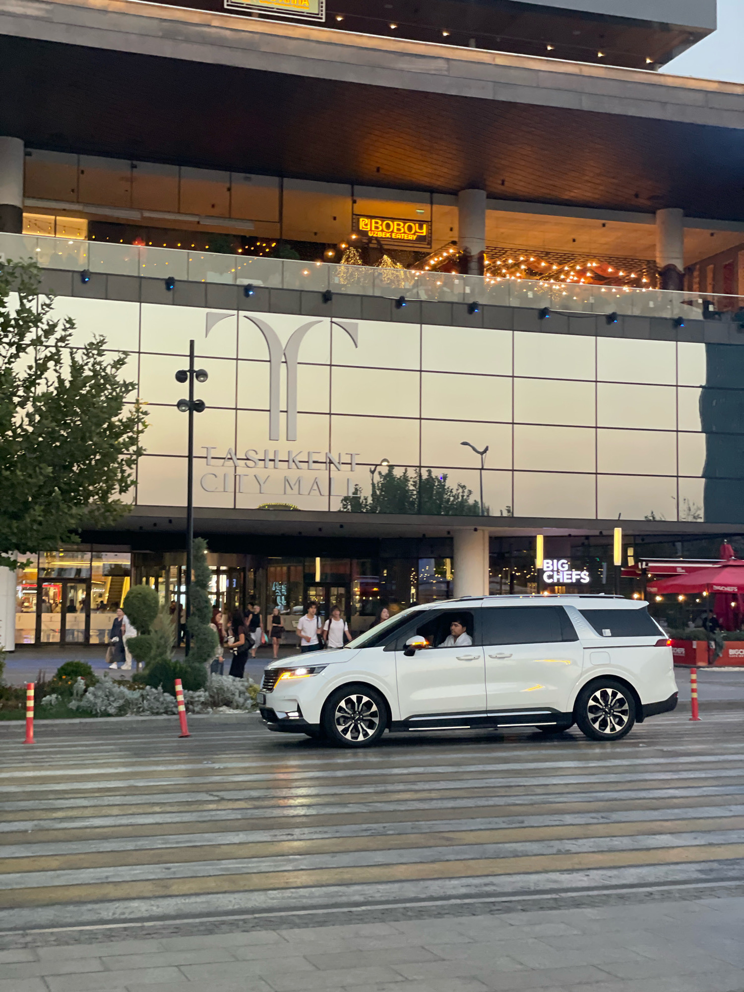 A white minivan crosses a crosswalk in front of the Tashkent City Mall in Tashkent, Uzbekistan. The mall features a large glass facade with the mall's logo and name, "TASHKENT CITY MALL," emblazoned on it. Several people are visible walking in front of the mall entrance, and there are trees and decorative plants in the foreground. The sky is overcast, and the scene appears to be during the day. The minivan has tinted windows, and one person is visible in the driver's seat. The crosswalk has yellow and white stripes, and there are orange and white bollards on either side of it. In the background, there are signs for "BOBOY UZBEK EATERY" and "BIG CHEFS." The overall impression is a modern urban setting.
