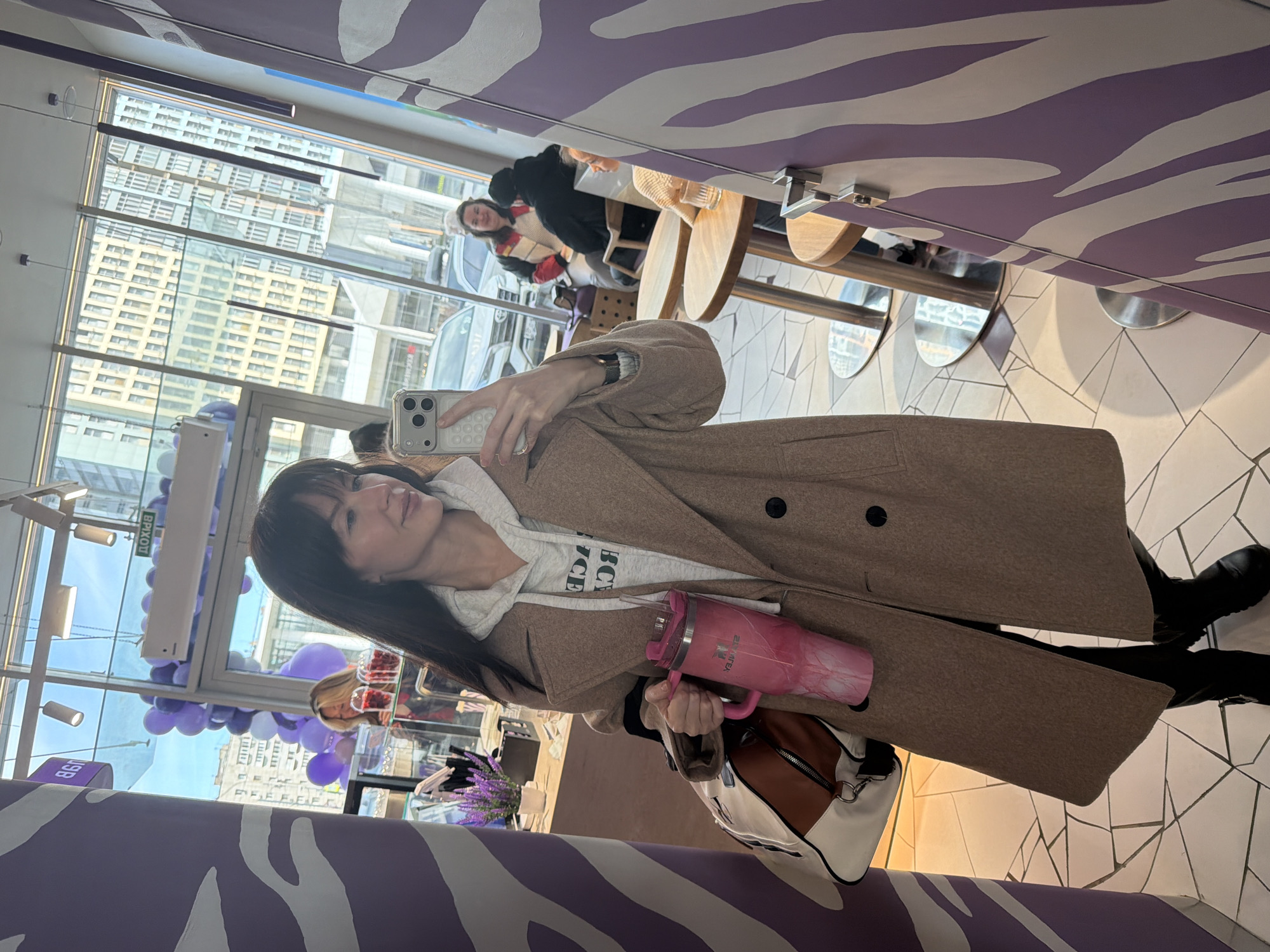 A woman stands in front of a purple and white zebra-striped wall, likely a design element of the interior, holding a phone in her right hand, presumably taking a photo of herself. She is wearing a long brown coat over a white hoodie, black leggings, and black boots. In her left hand, she holds a pink tumbler and a white and brown handbag. Behind her, a large window offers a view of a city with modern architecture, including tall buildings and cars on the street. The interior includes tables and chairs where other people are seated. The lighting is bright, likely due to the natural light streaming in from the large window. The scene is likely a café or restaurant. Based on visual clues like the exterior architecture visible through the windows and the interior design, the location is likely in Perm, Russia.