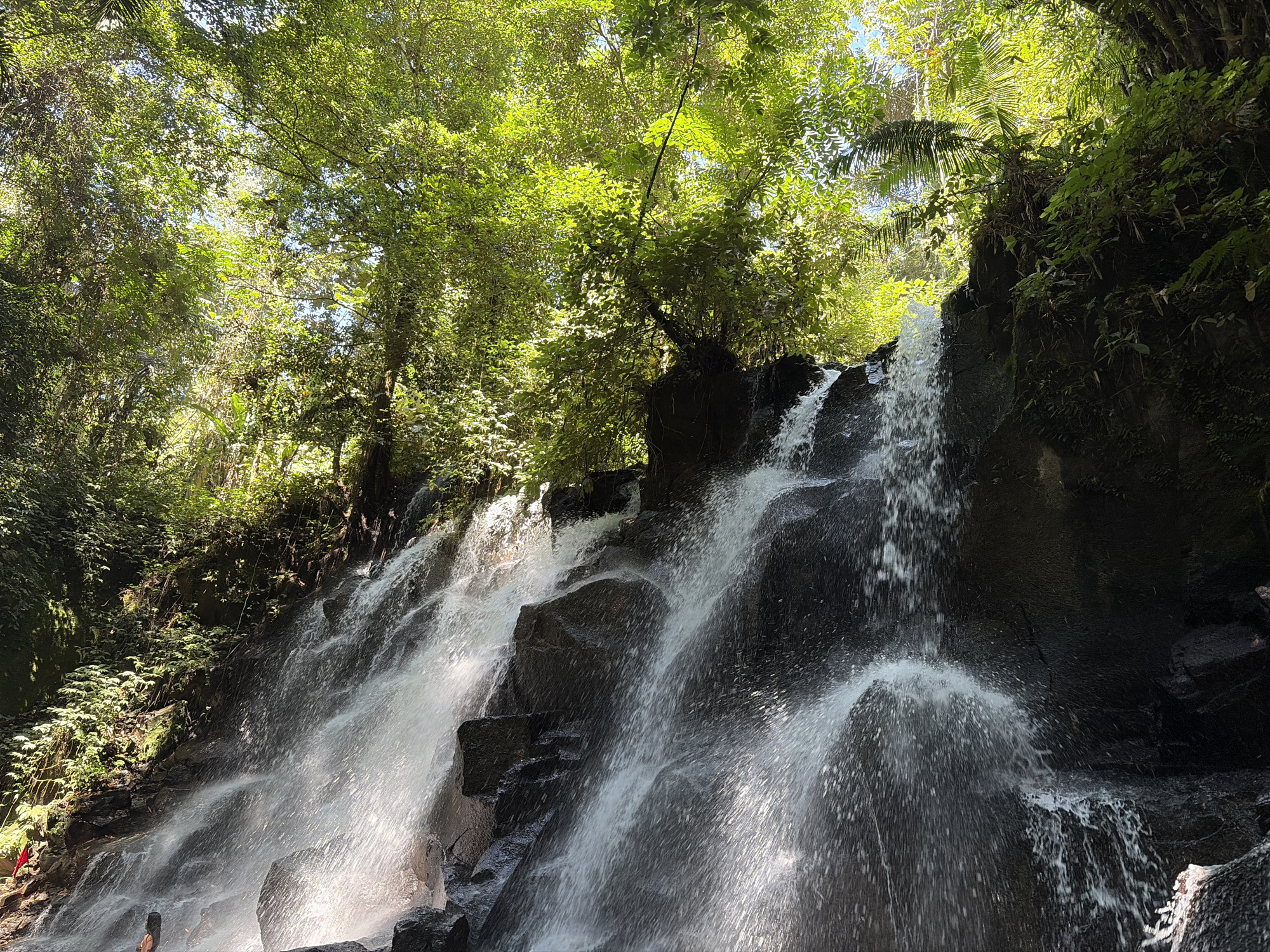 The image captures a cascading waterfall in a lush, tropical environment, likely in Gianyar, Indonesia, known for its natural beauty. The waterfall flows over dark, rugged rocks, with water appearing as white streaks and mist. Dense green foliage, including trees with sunlight filtering through their leaves, frames the scene from above and to the sides. The water spray creates a hazy effect, and the rocks are wet and dark, indicating the constant flow of water. In the lower left corner, a small section of a red flag is visible on the bank, and a person is partially visible standing near the base of the waterfall. The overall impression is one of a natural, vibrant, and serene landscape.