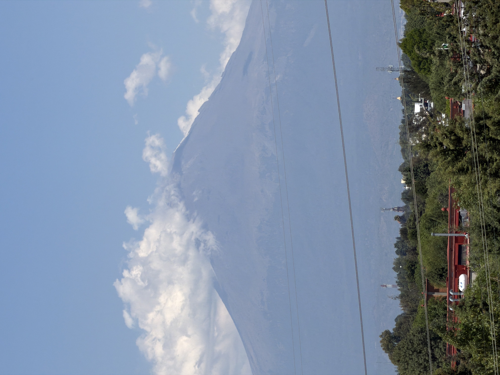 The image, when viewed in its correct upright orientation, presents a panoramic landscape with a massive volcano dominating the background under a bright, partly cloudy sky. The volcano, likely Popocatépetl given the location context of San Andrés Cholula, Mexico, features a snow or ice-capped peak, partially obscured by white cumulus clouds that also drift along its upper slopes. The atmosphere around the volcano appears somewhat hazy, giving its lower slopes a muted blue-grey tone.
Several thin utility lines stretch horizontally across the midground, intersecting the base of the distant volcano and the nearer landscape. Below these lines, the scene transitions into an urban or suburban area. Dense green foliage from numerous trees fills the midground and foreground, partially obscuring various buildings. A prominent structure with a reddish roof is visible on the right side of the foreground, nestled among the trees. In the distant midground, several communication towers, including one with distinct red and white sections, can be seen standing on a faraway ridge or hill.
The photograph was captured during bright daylight, indicated by the clear illumination and the vivid blue sky with scattered white clouds. No specific activity or event is taking place, and no people are visible. The overall impression is a serene, static capture of a volcanic landscape overlooking a developed area. There is no visible text in the image.