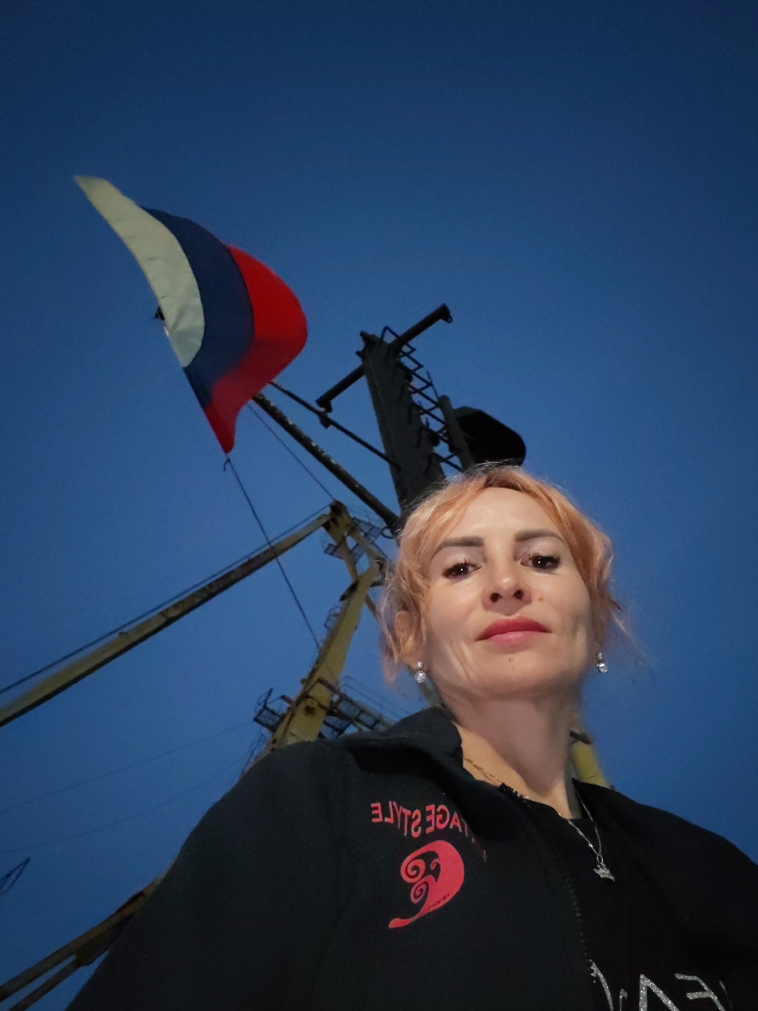 A medium shot captures a woman with auburn hair taking a selfie outdoors at dusk. She is wearing a black zip-up jacket with a red graphic design on the left side and a silver necklace. The Russian flag, colored white, blue, and red, is visible flying in the background against a clear blue sky. A portion of a yellow crane structure is also present in the background, suggesting an industrial or port setting. The location is stated to be in Bol'shie Mkhi, Russia.