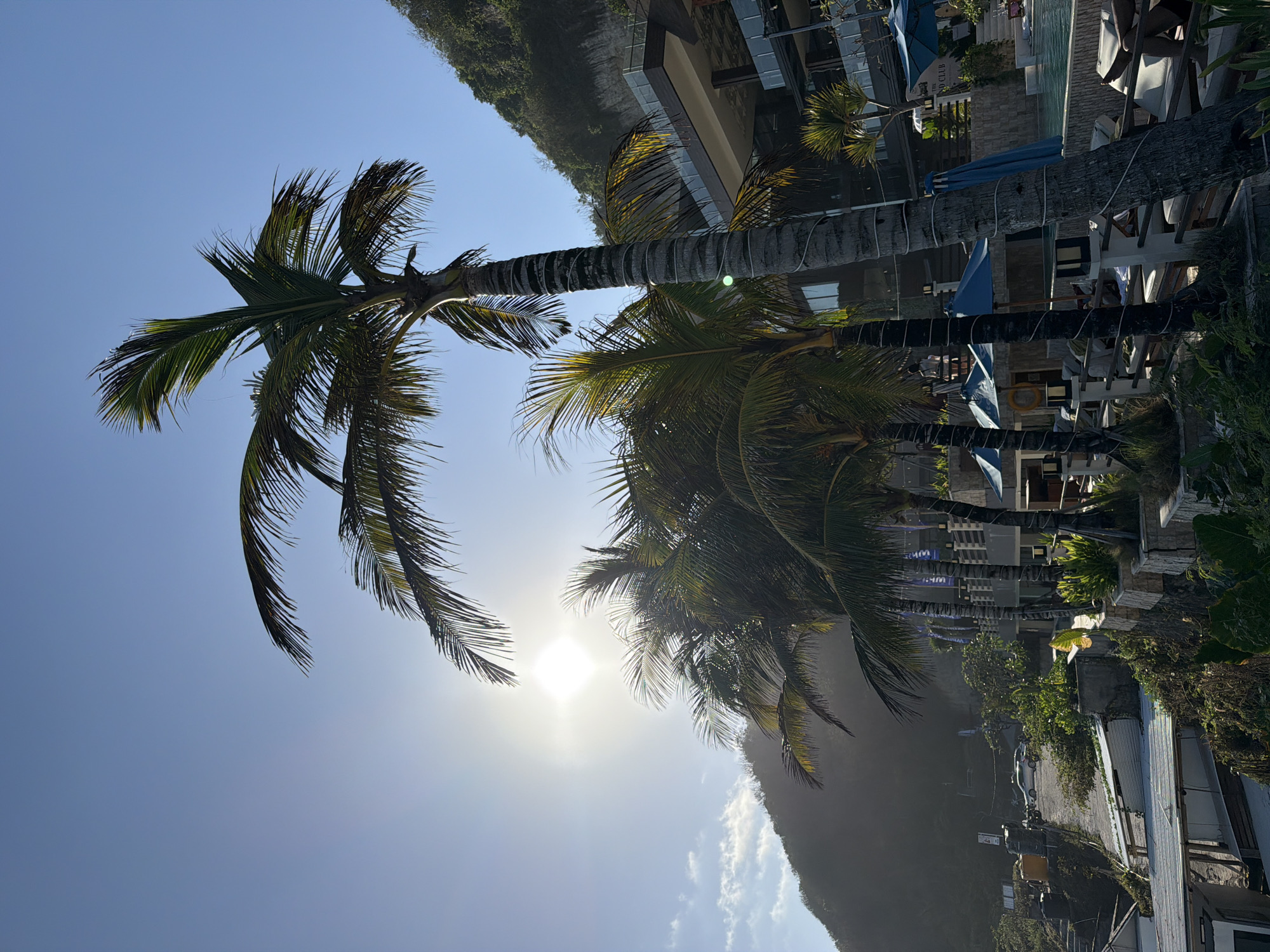 In Kuta Selatan, Indonesia, a resort scene unfolds under a bright, clear sky. Several tall palm trees with lush green fronds dominate the foreground, silhouetted against the sun. To the right, a multi-level resort building with a modern architectural design features balconies and glass windows. A swimming pool is visible, surrounded by lounge chairs and blue umbrellas. In the background, a steep, tree-covered hillside rises, partially obscured by the bright sunlight. A paved road winds up the hillside, with a white car visible on it. Some structures, possibly part of the resort or other buildings, are scattered along the slope, with metal-roofed structures in the lower left of the frame. The scene suggests a tropical vacation destination, bathed in the warm light of the sun.