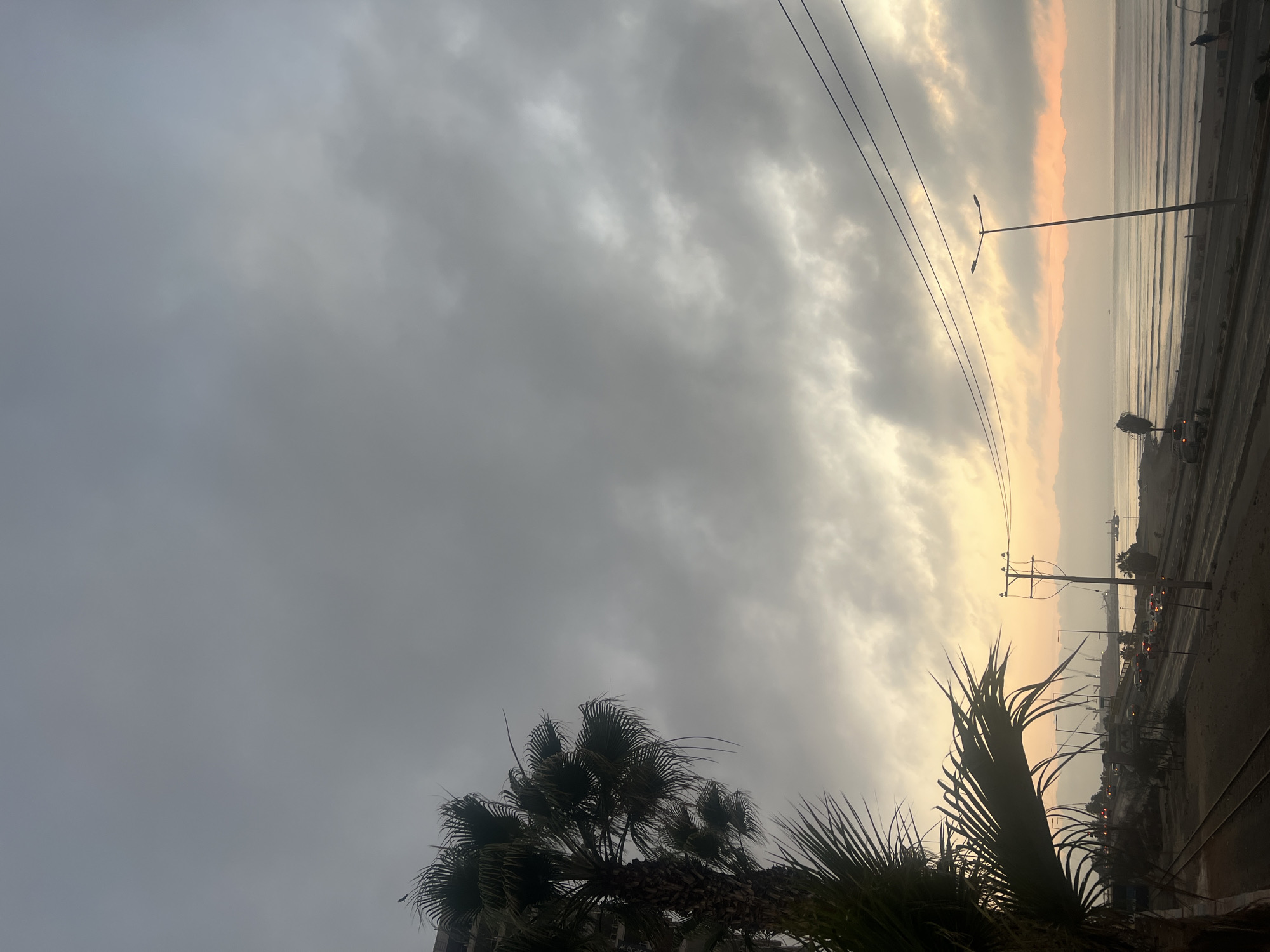 Arica, Chile: A landscape photograph shows a gray cloudy sky in the background. In the bottom center, there are palm trees. The top right section of the photograph shows the coast, buildings, and city streets. The sun is partially visible, creating a golden glow in the sky. Electrical lines run along the top portion of the image.