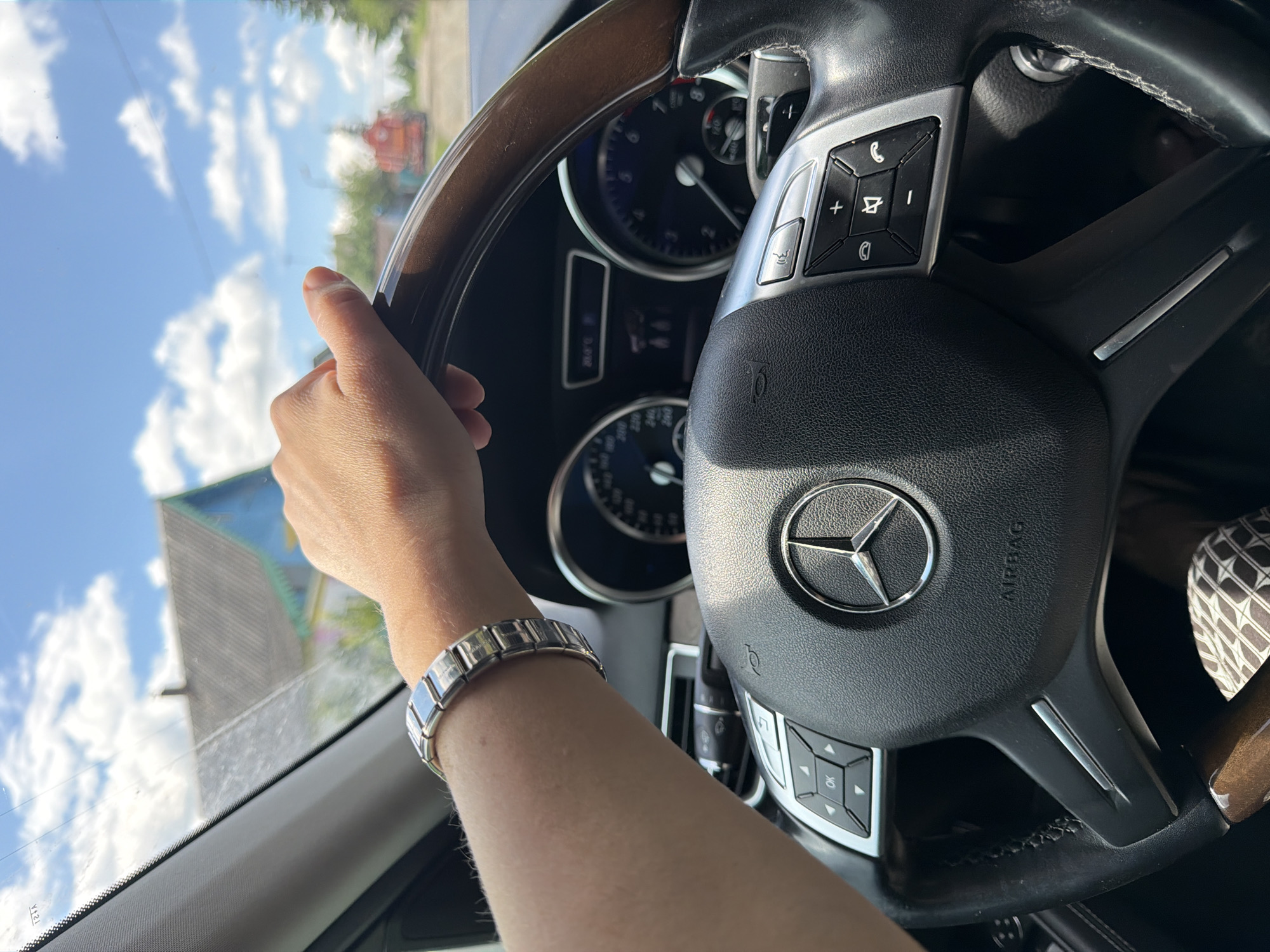 A first-person perspective shot captures a hand with a silver segmented bracelet resting on the steering wheel of a Mercedes-Benz, likely in Mezhevoy, Russia, given the presence of a distinctive blue and yellow house with a grey roof in the background and similar architecture in the distance, suggesting a rural or suburban setting. The sky is bright blue with scattered white clouds. The steering wheel is black leather with wood trim, and the car's dashboard features a Mercedes-Benz logo, air vents, and a digital display. The driver's arm is visible, extending from the left of the frame.