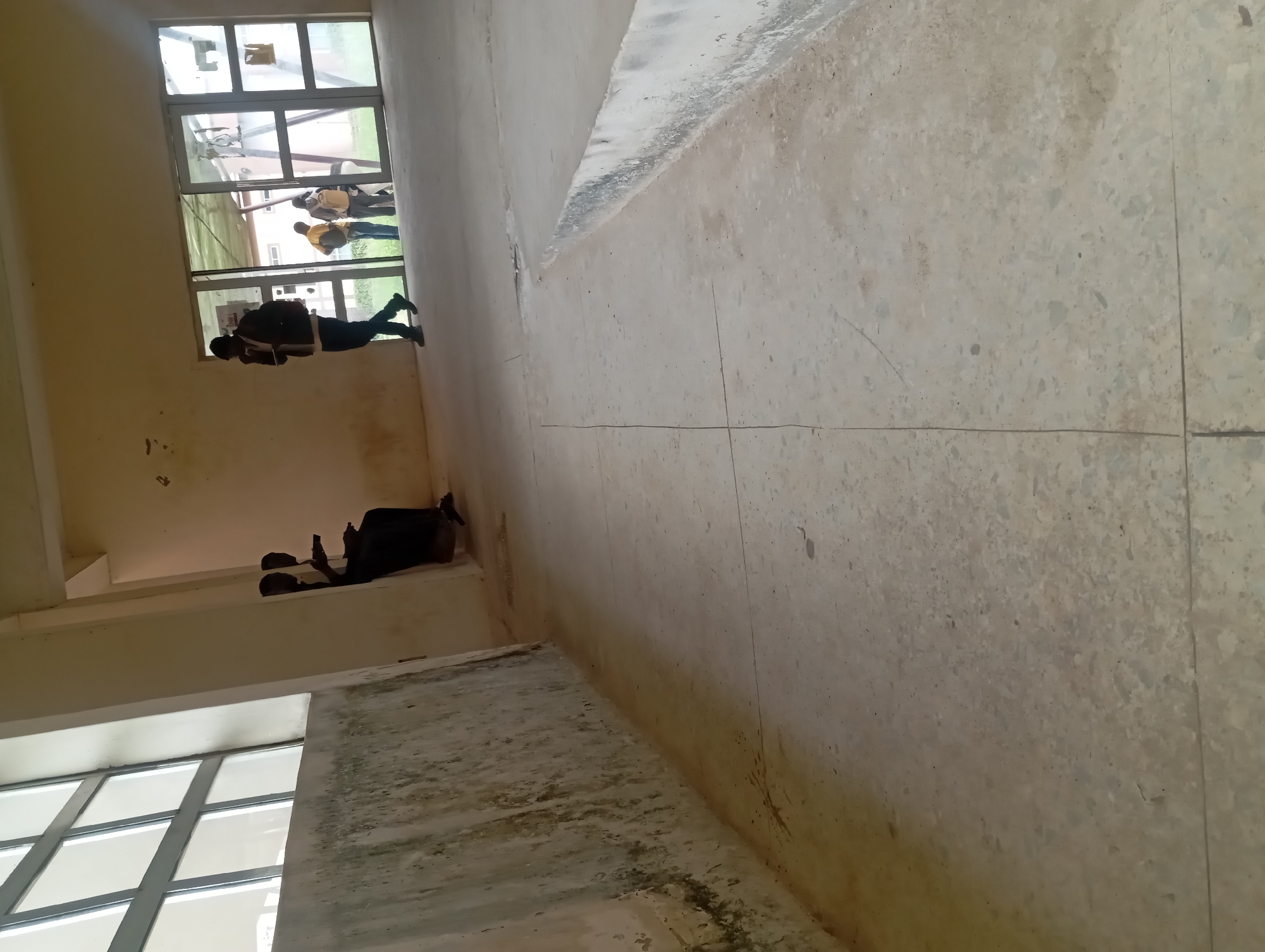 This is a photograph of an interior space, likely a hallway or open area within a building. The perspective is from a low angle, looking upwards and slightly towards the right, with the camera rotated. The dominant elements are the concrete floor, walls, and large windows.
There are several people visible. Near a large window on the left side of the image, a person is standing and looking at a phone. Further down the hallway, also near a window, two people are seated or reclining on a raised platform. One of them is also looking at a phone. Through the large windows in the background, more people can be seen, some appearing to be standing and talking. These individuals are wearing casual clothing, including t-shirts and backpacks, suggesting they are students or staff in an educational or public institution.
The scene appears to be well-lit, likely due to the large windows. The time of day cannot be determined with certainty, but the ambient light suggests it is daytime. The concrete floor has a somewhat worn appearance with some discoloration and cracks. The walls are painted a light color and show some signs of age or staining. The overall impression is of a functional, possibly slightly neglected, interior space.
There is some visible text on a white sign inside the window on the left, but it is too small and blurry to be legible.