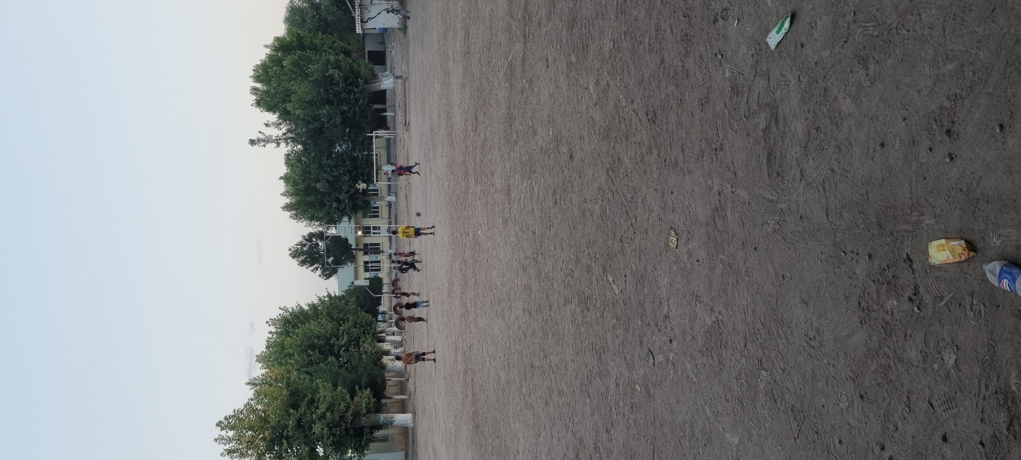 In Хаваскор, Tajikistan, an outdoor scene unfolds on a dusty, dirt field where a group of children are engaged in a game of soccer. The sky above is a pale, hazy blue, suggesting either early morning or late afternoon light. The field itself is largely barren, with tire tracks and scattered debris visible on the surface, including a discarded Pepsi bottle and a crumpled yellow and red wrapper.
To the left and in the background, a line of trees with full green foliage provides a natural border. Behind the trees, several buildings are visible, appearing to be low-rise structures with light-colored walls and dark-trimmed windows. One building in the mid-ground has a slightly more prominent facade with several windows and a light-colored roof. In the far background, a larger, lighter-colored building with a visible roofline is present.
The children, numbering approximately ten, are scattered across the field. Some are actively playing soccer, with one boy in a yellow shirt with the number "7" clearly visible, and another in a blue and red soccer uniform. Others are standing or running. There are also a few scattered tires on the field, possibly used as markers or obstacles. The overall impression is a casual, community sporting activity taking place in a somewhat unkempt, rural or suburban setting.