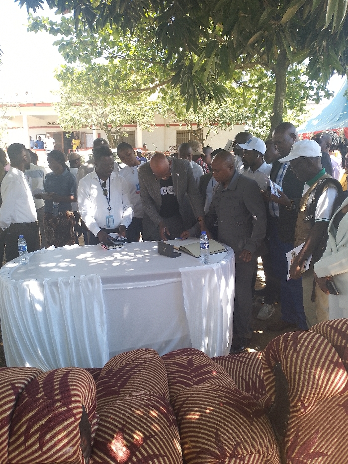 A group of 15 men are gathered around a white table covered in a white tablecloth in Bunia, Democratic Republic of the Congo, located at coordinates 1.556 latitude, 30.244 longitude. The men are dressed in various attire, some wearing suits and ties, others in shirts and slacks, and some in more casual clothing. They are gathered around a table with two bottles of water on it, and a man in a black suit is signing a document. The scene appears to be an official ceremony or meeting, with the men likely discussing or signing important documents. The environment is outdoors, with a large tree casting shade on the scene. There are striped pillows on the ground in the foreground.