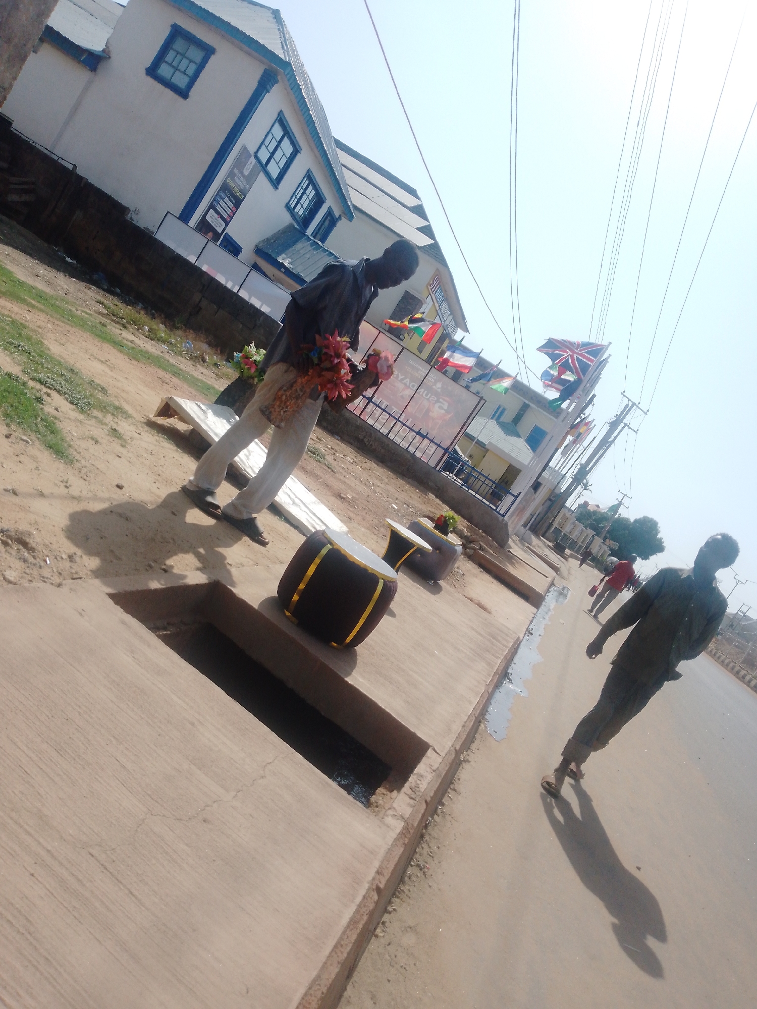 The scene is set in Sabon Tasha, Nigeria. A man stands on the sidewalk, holding a bouquet of artificial flowers. A drum rests near a drainage grate. Another man is walking on the road. In the background is a building adorned with international flags, including the Union Jack. Overhead power lines crisscross the sky.