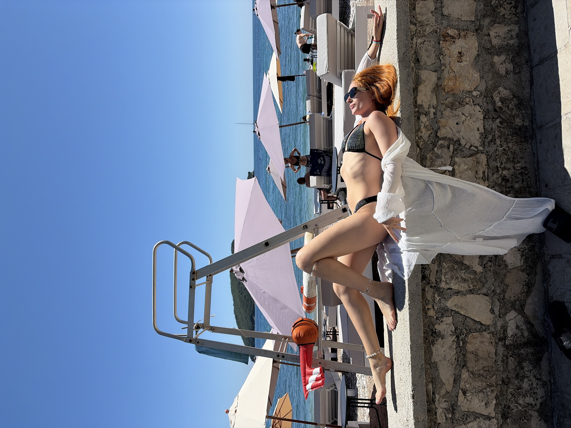An outdoor, sun-drenched scene captures a woman relaxing on a stone ledge at a beach club in Budva, Montenegro. The main subject, a woman with reddish-blonde hair pulled back, is lying on her back, wearing dark sunglasses, a dark sequined bikini top and bottom, and a sheer white cover-up draped over her body. Her right arm is extended above her head, resting on the textured stone wall behind her, while her left arm lies across her midsection. She has a pearl anklet on her left foot, which is crossed over her right leg.
In the mid-ground, to the left of the woman, the clear blue sea is visible, dotted with several light-colored beach umbrellas (some pinkish-white, others beige-white) and white lounge chairs. Other individuals are present in the background, some relaxing on chairs, one standing in the water, and another near the umbrellas. A metal lifeguard stand or diving platform structure is prominent on the left side of the frame, featuring an orange and red inflatable floatation device attached to it. The sky above is a vibrant, cloudless blue, indicating clear, sunny weather, likely mid-day due to the bright light and strong shadows. The stone surface she is resting on and the adjacent textured stone wall provide a rugged contrast to the soft beach elements. A single black shoe or slipper is visible near her feet on the stone ground. There is no clearly visible or readable text in the image.