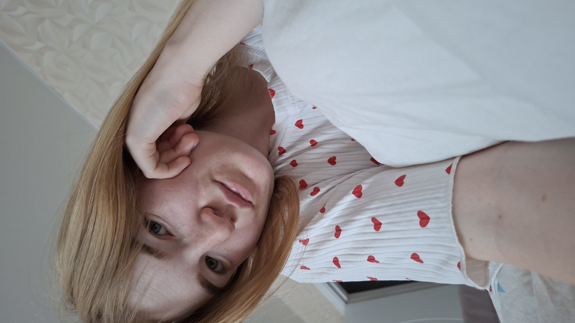 A young woman is reclining, looking directly at the camera. She has light skin, light brown hair, and is wearing a white shirt with red heart patterns. Her left hand is positioned near her ear. The location is Chaikovsky, Russia, discernible from metadata not included in the image. The environment appears to be indoors, based on the wallpaper and lighting.