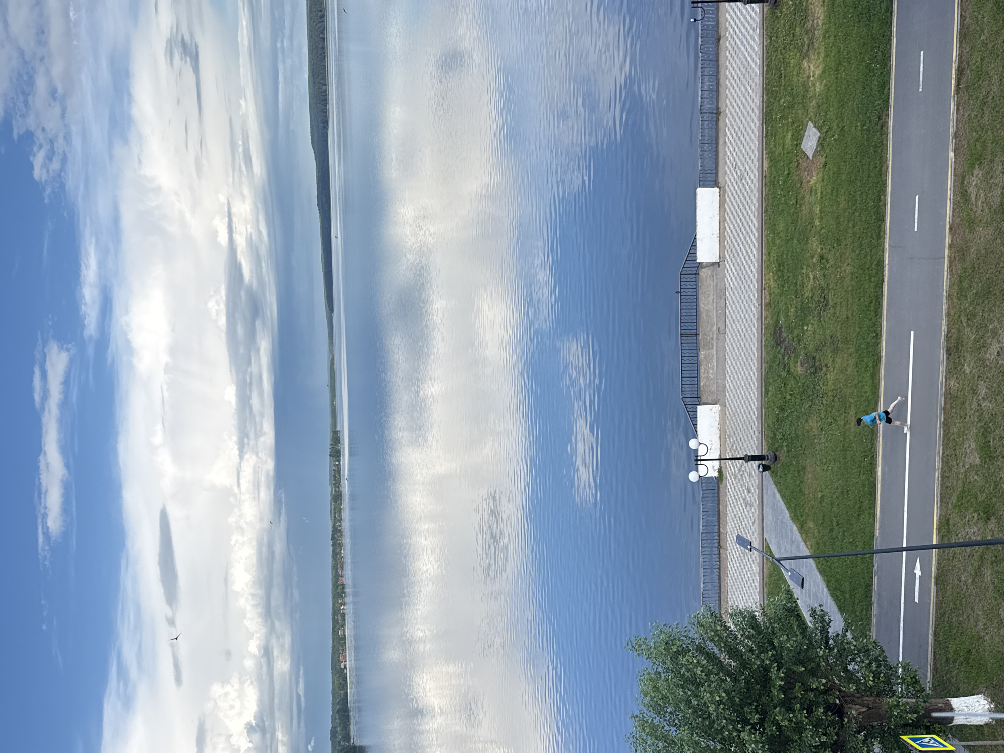 The image, taken in Votkinsk, Russia, shows an elevated view of a waterfront scene. A wide body of water (presumably a lake or river) occupies the left half of the frame, reflecting the blue sky and clouds. The right half depicts a paved walkway bordered by a strip of grass. A pedestrian is visible jogging along a marked pathway adjacent to the grass. Streetlights are spaced along the walkway. A portion of a tree is in the foreground. At the bottom corner is a pedestrian crosswalk sign. The scene is taken in daylight with clear weather.