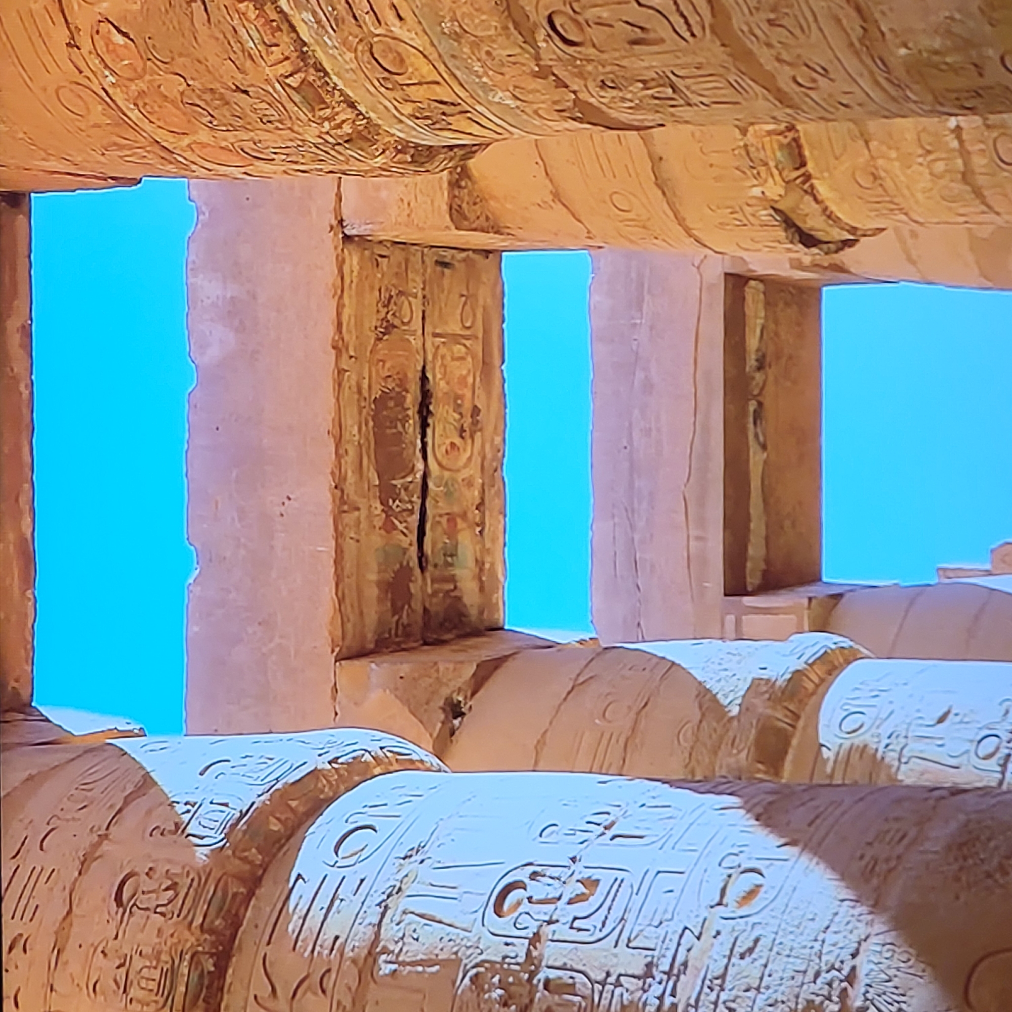 The image depicts architectural elements of ancient Egyptian construction, specifically cylindrical columns and lintels adorned with hieroglyphics. The columns are arranged in a row, with sunlight creating shadows that emphasize the carved details. The lintels above show more hieroglyphic inscriptions. Rectangular openings provide a view of a clear, bright blue sky. The scene suggests a temple or other monumental structure from ancient Egypt. The image cannot be located in Yerevan, Armenia; instead, it must be located at an ancient Egyptian temple site.
