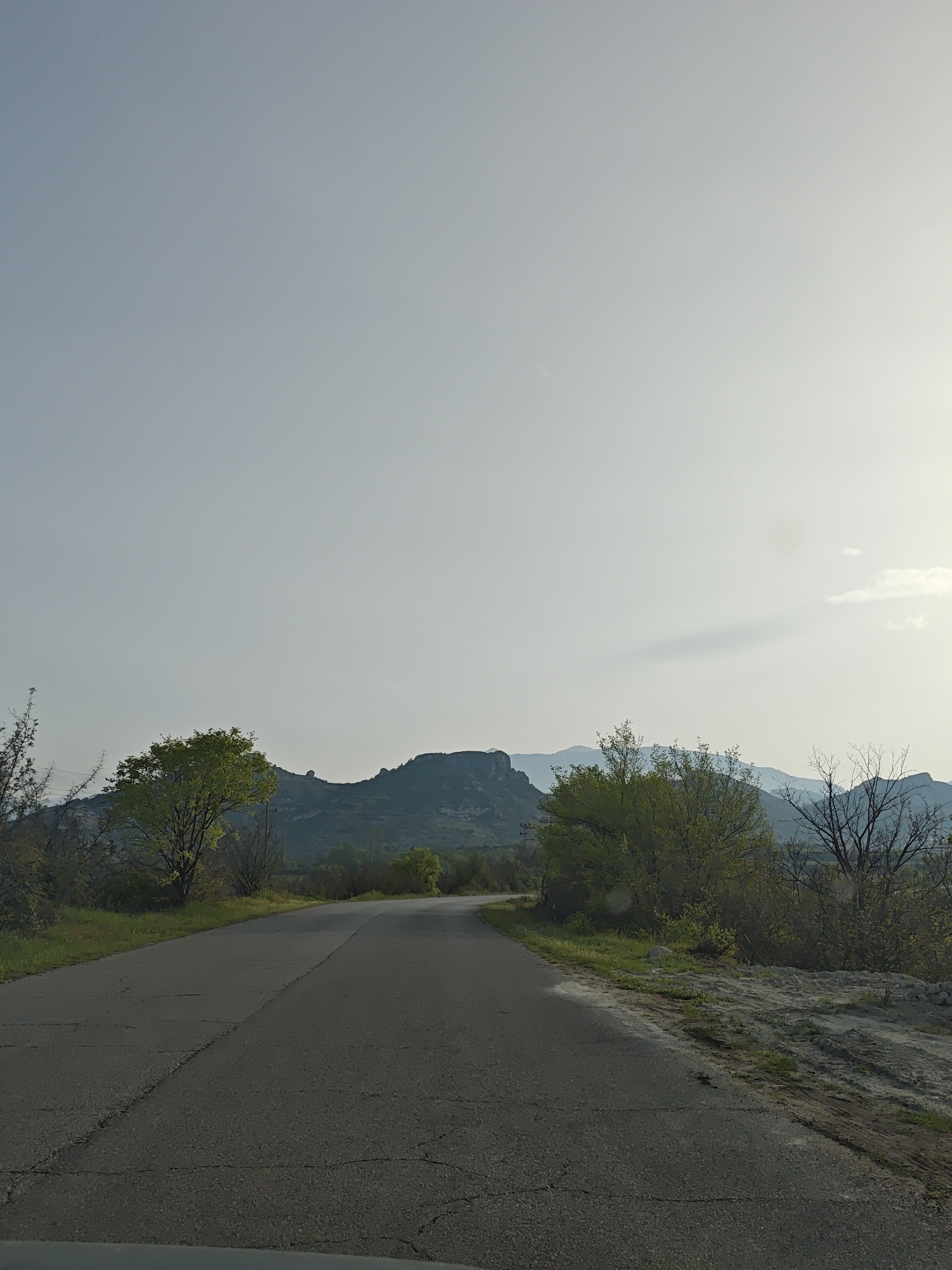 A paved road stretches into the distance, curving slightly to the left. The road surface is dark and shows signs of wear and cracking. On either side of the road, there is vegetation, including trees and shrubs with sparse foliage, suggesting either early spring or a dry environment.
In the background, a range of hills is visible, with one prominent hill standing out. The sky is overcast and light in color. No people are visible in the scene. The setting appears to be a rural or semi-rural area. The location is Unknown, Unknown.