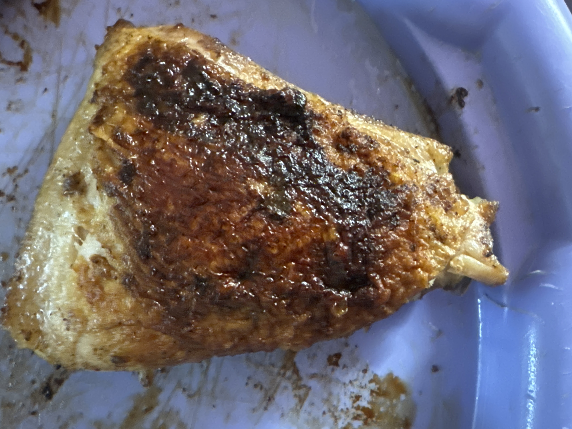 A close-up, top-down shot shows a piece of fried chicken on a light purple plastic plate. The chicken is triangular with a golden-brown, slightly charred crust. The skin is glistening with oil, and darker, caramelized bits are visible. The plate shows some smudges and small dark specks around the chicken, indicating previous food contact. The background is out of focus, revealing the light purple texture of the plate and some indistinct brown residue. The image appears to be taken indoors. There is no information available about the location in the city of Bohunove, Ukraine.