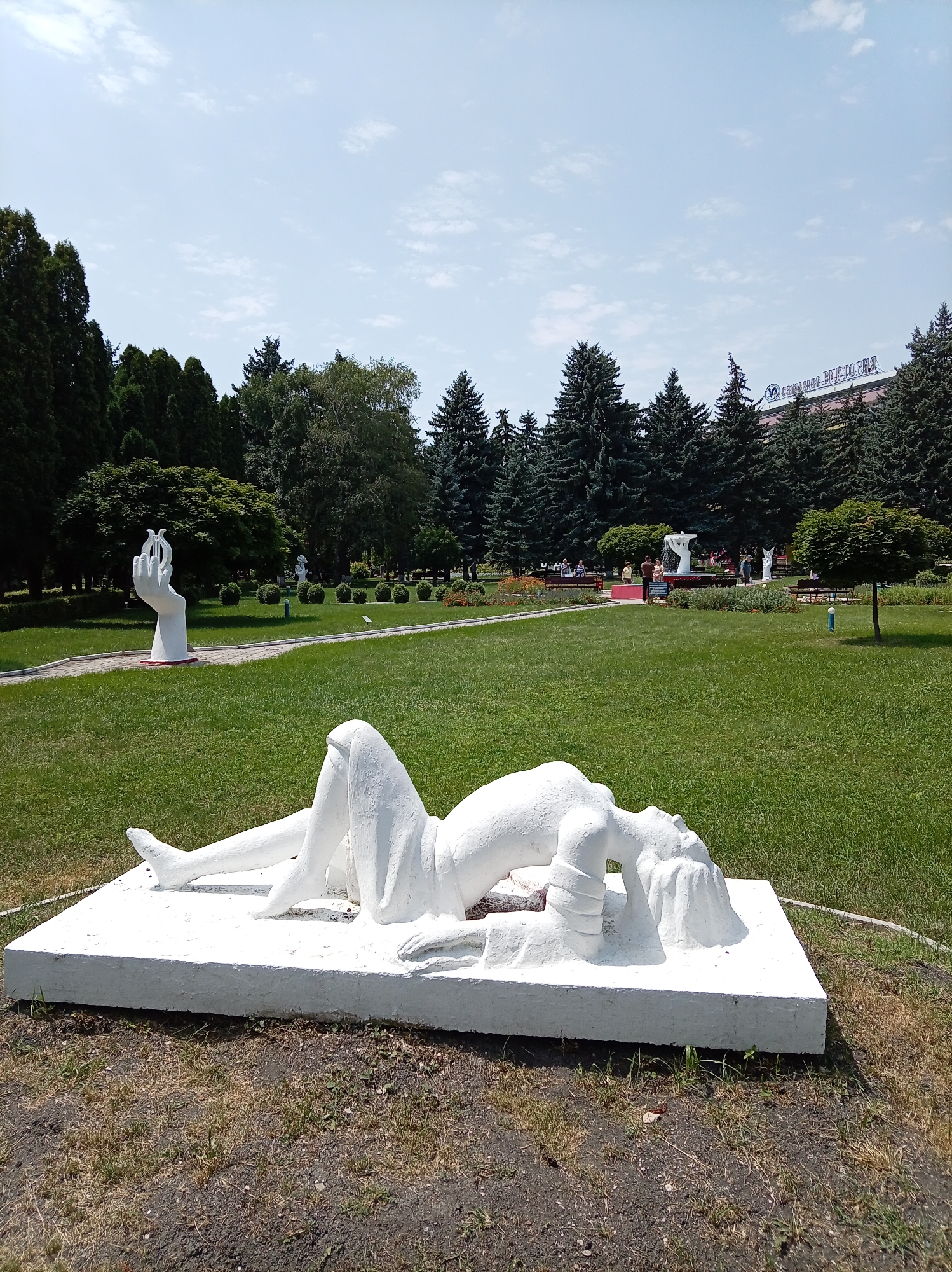 The image depicts a white statue of a woman in a reclining, arched pose on a rectangular concrete base in the foreground. The statue is situated in a park-like setting with lush green grass. In the background, there are numerous tall evergreen trees. Further back, a building with a distinctive sign is visible, partially obscured by trees. There are also other sculptures, benches, and a fountain in the mid-ground, with a few people milling about. This scene is located in the central park of Yessentuki, Russia, specifically near the location of the monument to Vladimir Mayakovsky, which is referenced by the building sign indicating "Mayakovsky". The weather appears to be clear and sunny.