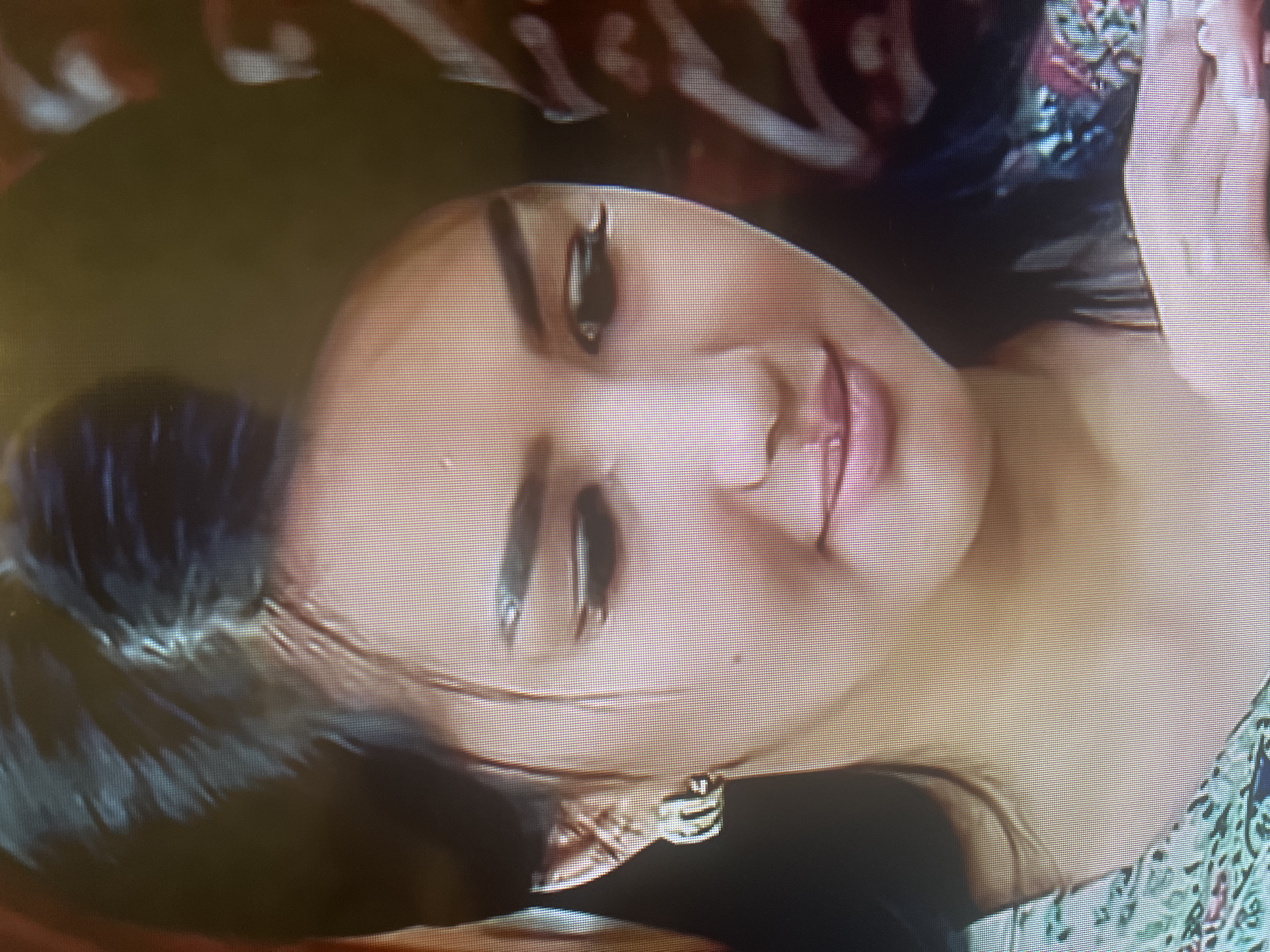 The image is a close-up, slightly pixelated shot of a young woman's face. She has dark hair pulled back, accentuating her facial features. She is wearing a silver earring on her left ear, which appears to be in the shape of the letter 'M' or a stylized elephant. Her eyes are lined with dark makeup, and her eyebrows are well-defined. Her lips are slightly parted, and she has a subtle smile. The lighting suggests an indoor setting, possibly a dimly lit room. The background is out of focus, showing a mix of dark and patterned textures, suggesting fabric or upholstery. The woman's clothing is visible at the bottom left of the frame, appearing to be a patterned fabric with green and pink floral motifs. The image does not contain any elements that would allow for determining its location in Osh City, Kyrgyzstan.