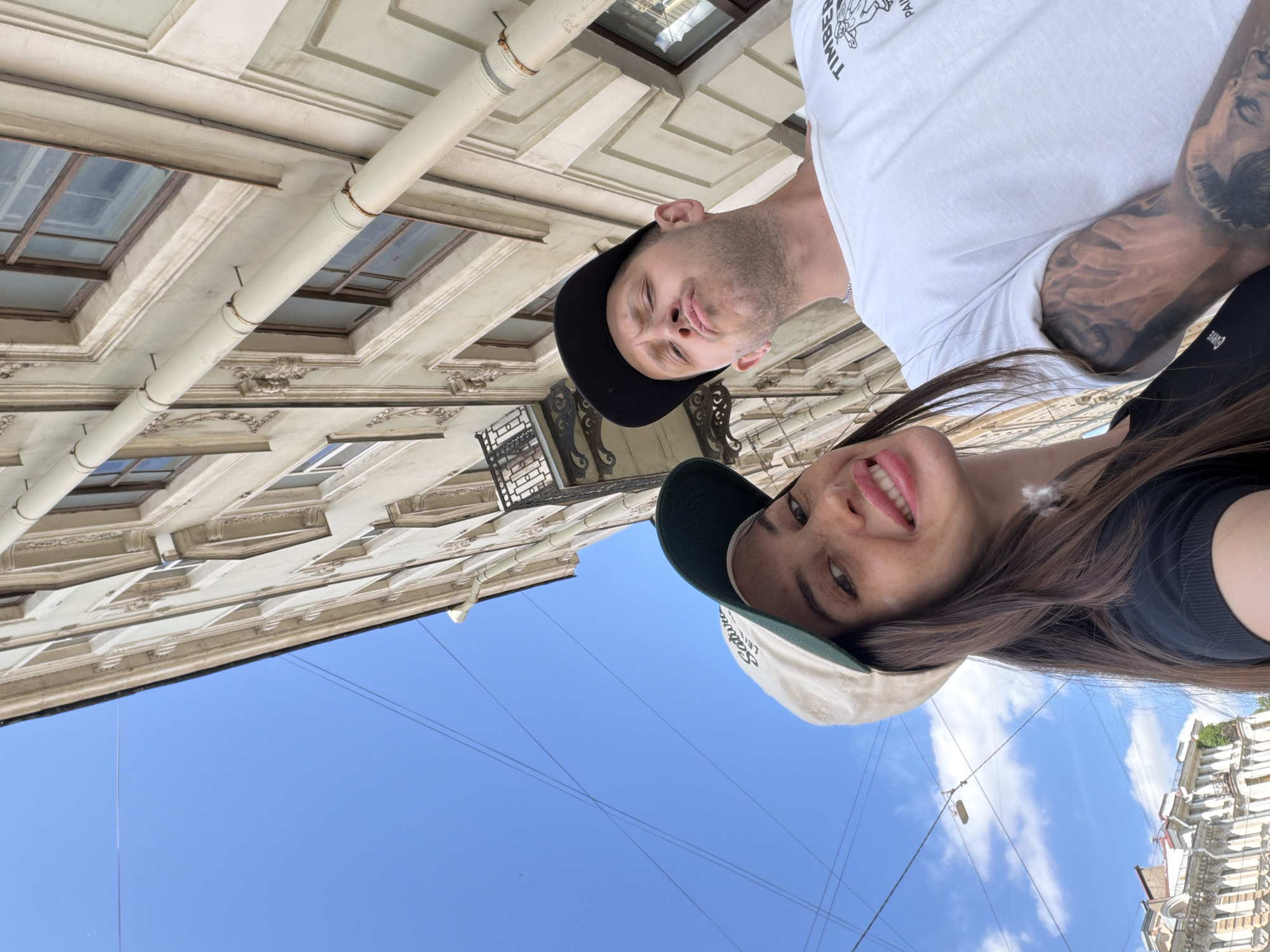 An outdoor, low-angle, medium shot captures a young white couple posing for a selfie with an ornate, cream-colored building in the background, beneath a clear blue sky. The woman, on the left, wears a black t-shirt and a cream-colored baseball cap with a green brim and embroidered text. She looks at the camera with a wide smile, revealing her teeth. She has long, dark brown hair and freckles across her nose and cheeks. The man, on the right, wears a white t-shirt and a black baseball cap. He makes a pouty face towards the camera. He has a buzz cut and a light beard stubble. The building features a drainpipe running vertically along its facade, with intricate architectural details and balconies. Power lines are visible against the sky. The city of Saint Petersburg, Russia, is identified as the location.