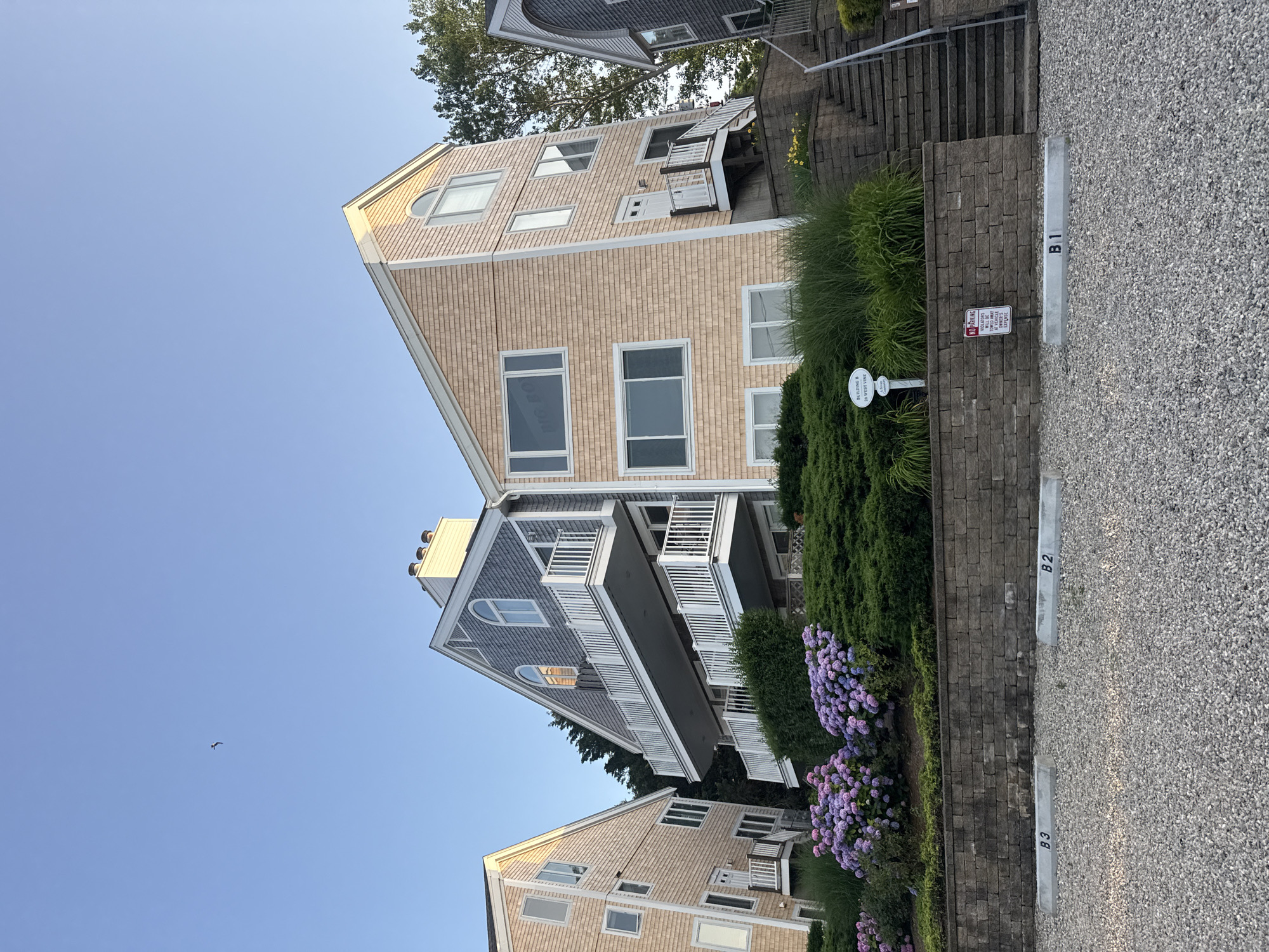 In Provincetown, Massachusetts, USA, a multi-story residential building with light peach-colored shingle siding is photographed at a low angle, showcasing its distinctive architecture. The building features multiple balconies with white railings and several windows, including a prominent arched window at the top. Adjacent to it is a building with gray shingle siding. The foreground is a gravel parking lot with concrete parking space markers labeled B1, B2, and B3. Behind the parking lot, a low stone wall supports a terraced garden area with lush green bushes and vibrant purple hydrangeas. A sign on a post in the garden reads "BUILDING B 26 WEST VINE." A red and white "NO PARKING" sign is also visible on the stone wall, indicating that violators will be towed. The sky above is clear and blue, with a single bird in flight. The scene is illuminated by natural light, suggesting it is either early morning or late afternoon.