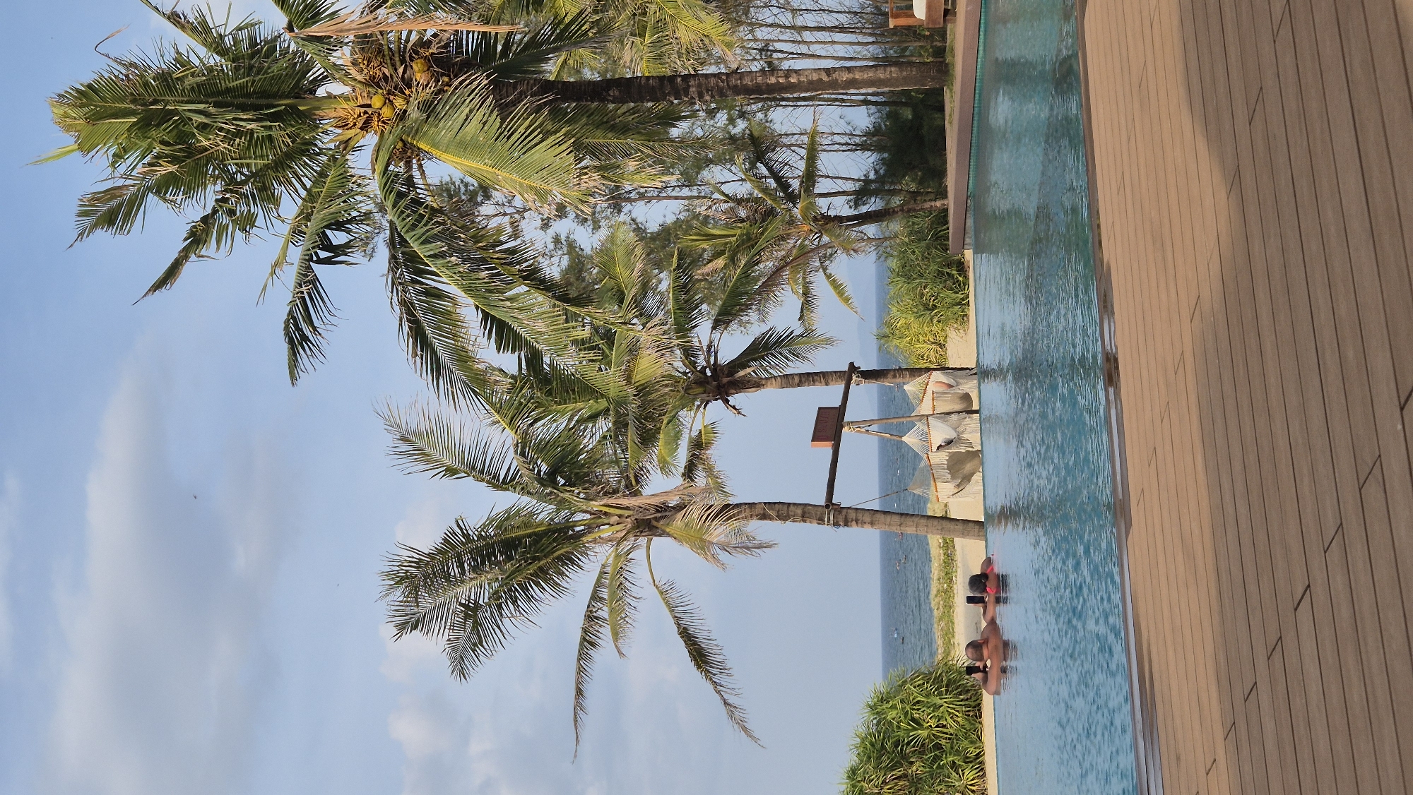 An aerial view captures a resort pool area in Hoi An, Vietnam, with the ocean in the background. In the foreground, a wooden deck leads to a vibrant blue swimming pool. Two individuals are visible in the pool, relaxing on inflatable tubes. Beyond the pool, a sandy beach is lined with lush green palm trees and tropical foliage. A hammock structure is suspended between two palm trees, adding a touch of leisure to the scene. The ocean stretches out to the horizon under a clear sky with scattered clouds. The overall impression is one of tropical serenity and a luxurious vacation experience.