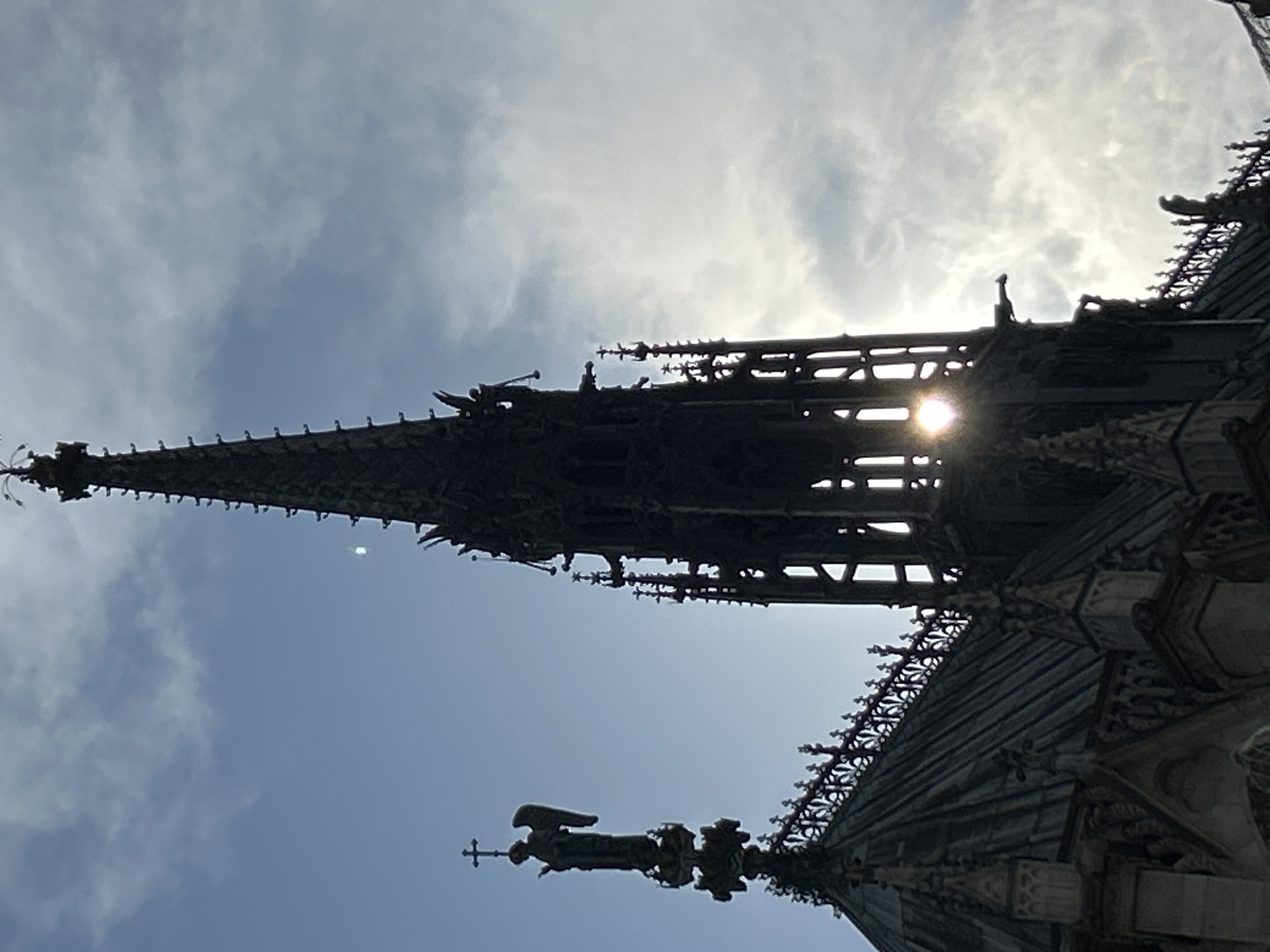 The image shows a close-up, low-angle view of the spire and rooftop of Notre Dame Cathedral in Paris, France. The spire, a dark silhouette against a bright sky, is intricately detailed with Gothic architecture. Gargoyles are visible perched on ledges, and the sun glints through the latticework of the spire. To the left, an angel statue stands atop a smaller pinnacle, holding a cross. The sky is a mix of blue and cloudy, with the sun creating a lens flare. The foreground shows the dark, sloped roof of the cathedral, also adorned with decorative architectural elements. The overall impression is one of grandeur and architectural complexity, emphasizing the Gothic style of the cathedral.