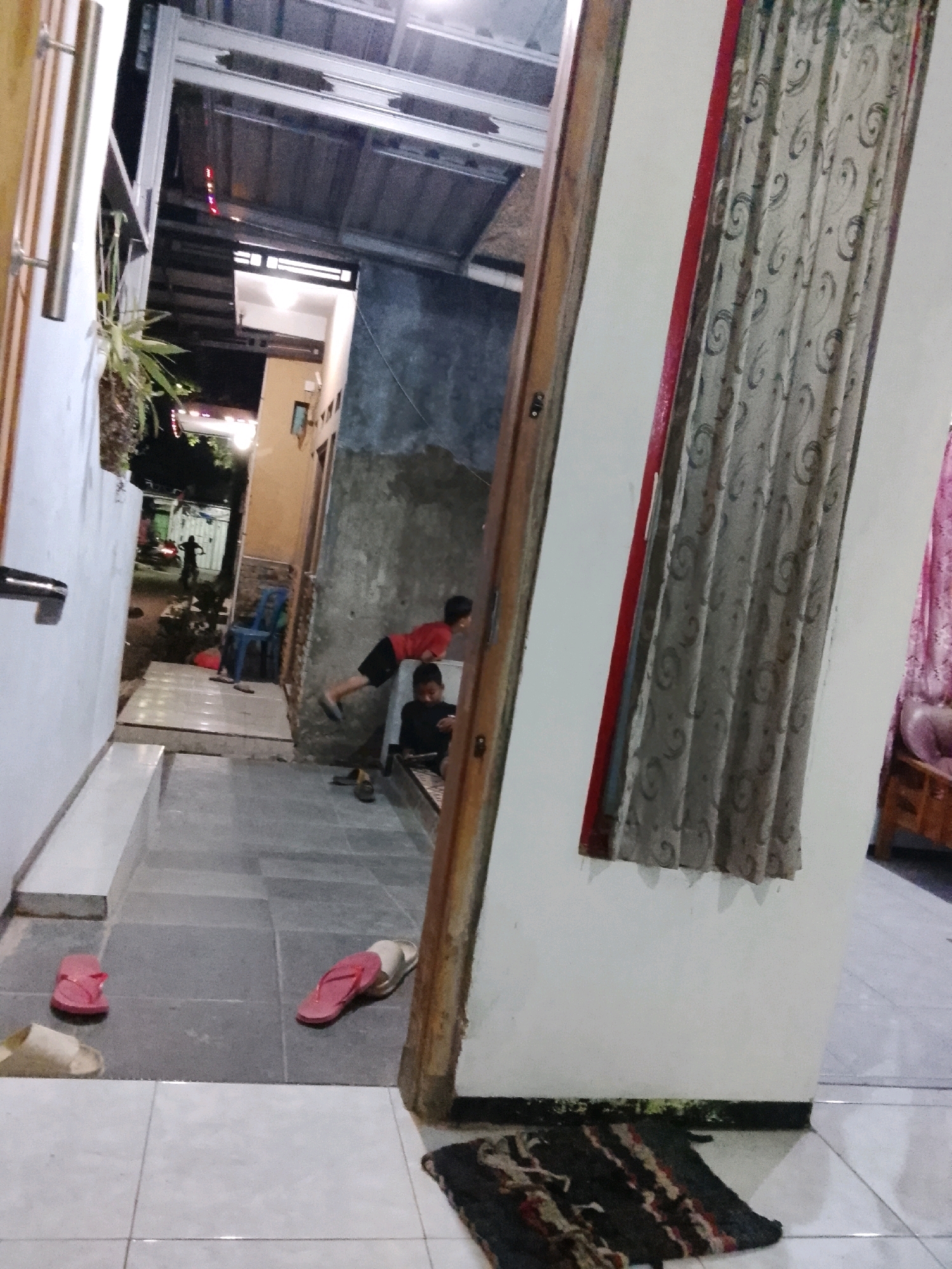 The image depicts an outdoor narrow walkway between two buildings, likely in the city of Ujungrusi, Indonesia. The walkway is paved with stone tiles and the ground level is uneven. On the left, a white wall with a metal handrail runs along the walkway, partially obscured by a hanging plant. On the right, a beige wall is prominent, featuring a curtain with a swirling pattern hung in front of a window.
Two young boys are visible in the scene. One boy, wearing a red t-shirt and black shorts, is in mid-air, leaning over as if jumping or being held. He is positioned on a slightly elevated section of the walkway. Below him, a second boy, dressed in black, sits on a mat or cushion.
Several pairs of flip-flops are scattered on the walkway: a pair of pink flip-flops near the bottom left, and another pair, one pink and one beige, near the center left. A dark-colored mat with red and beige stripes lies on the tiled floor in the foreground on the right.
Further back in the walkway, there is a blue plastic chair, and a glimpse of a motorcycle. The background shows a residential area with some lights visible, indicating it might be evening or night. The overall scene suggests a casual, everyday environment within a residential compound.