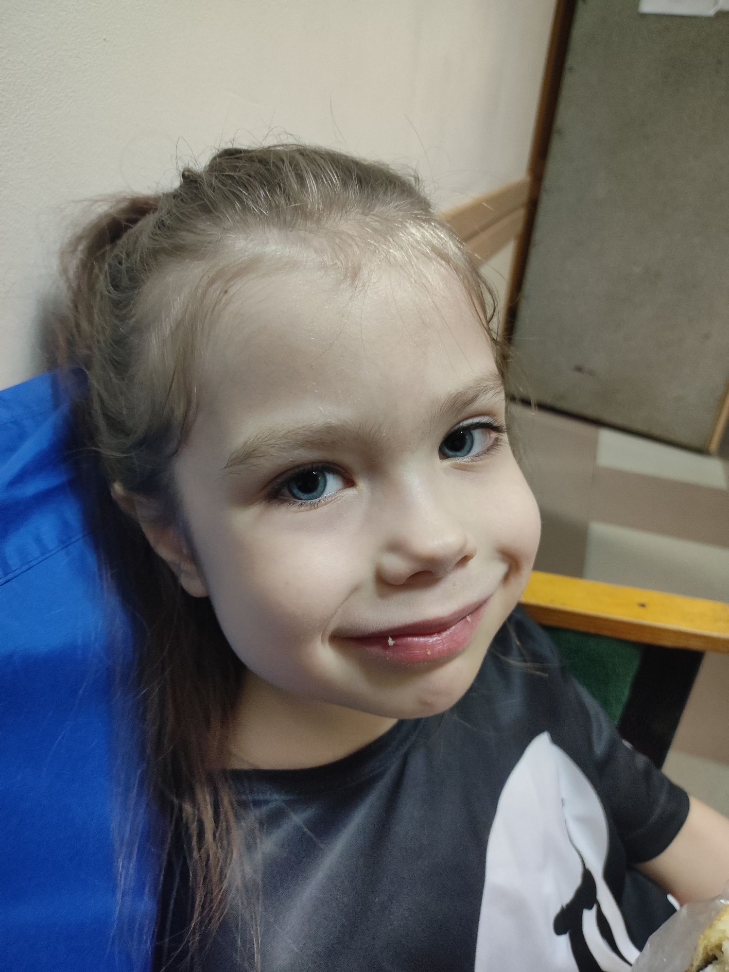 A close-up, slightly upward-angled shot features a young girl with fair skin, light brown hair pulled back into a ponytail, and striking blue eyes. She has a gentle smile on her face, and some crumbs are visible on her lips, suggesting she may have recently finished eating.
She is sitting on what appears to be a chair with a blue cushion. She's wearing a dark t-shirt with a white graphic on the front. The background is indistinct but reveals a cream-colored wall on the left and a dark wood panel along the right side of the frame, the interior of a building.
There is no indication of Votkinsk, Russia in the provided image.