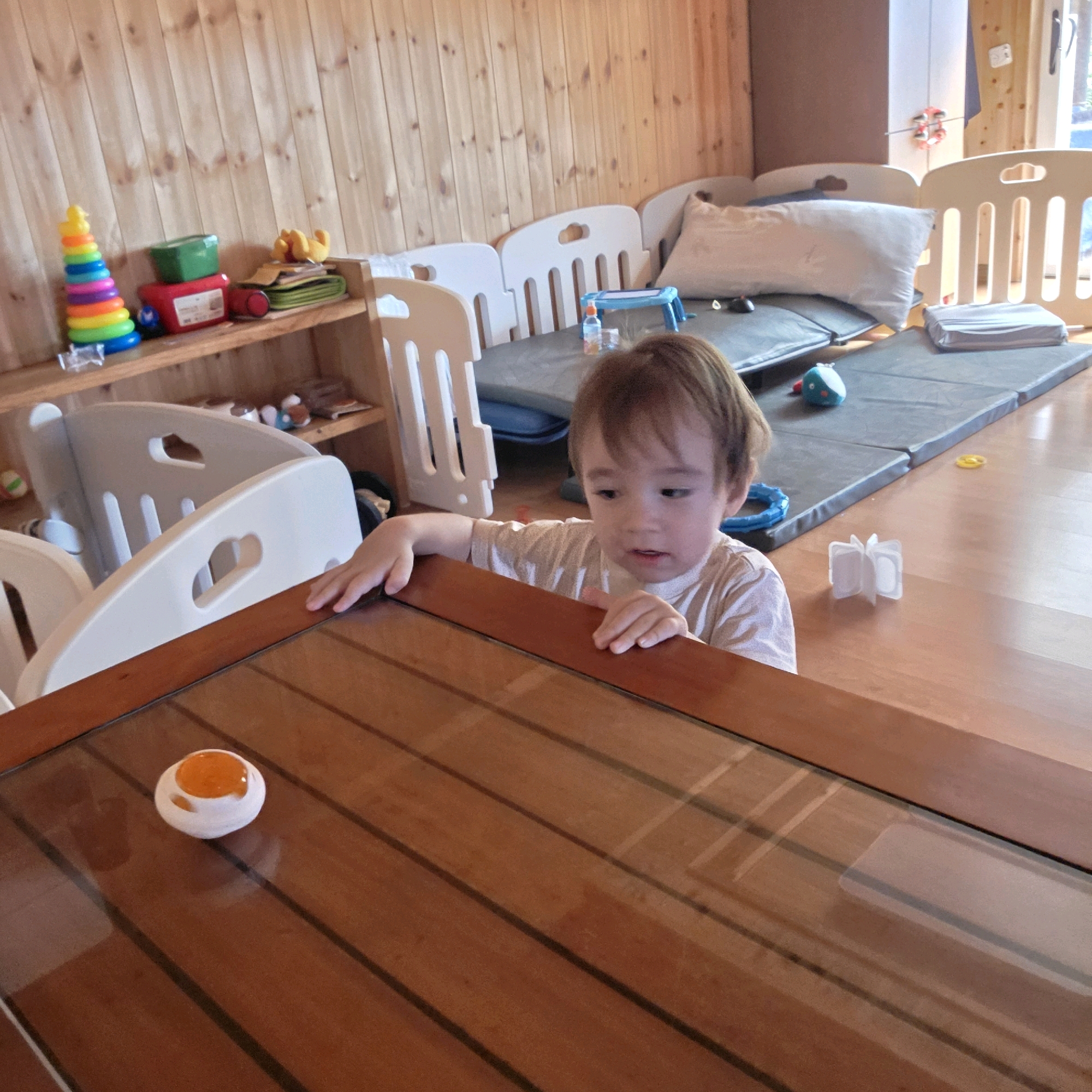 The image depicts an indoor scene, likely a playroom or daycare, as suggested by the presence of children's toys and play equipment. A young child with light brown hair is leaning on a wooden table, their hands resting on its surface. The child is wearing a light-colored t-shirt. The table has a reflective, possibly glass, top, on which a small, round object with an orange top and white base is placed. In the background, a wooden wall and shelves filled with toys are visible, including a stacking ring toy, a red toy car, and a green container. Further in the background, play mats and children's safety barriers are arranged, indicating a dedicated play area. There are no explicit indicators to confirm the location as Dunnae-myeon, South Korea.