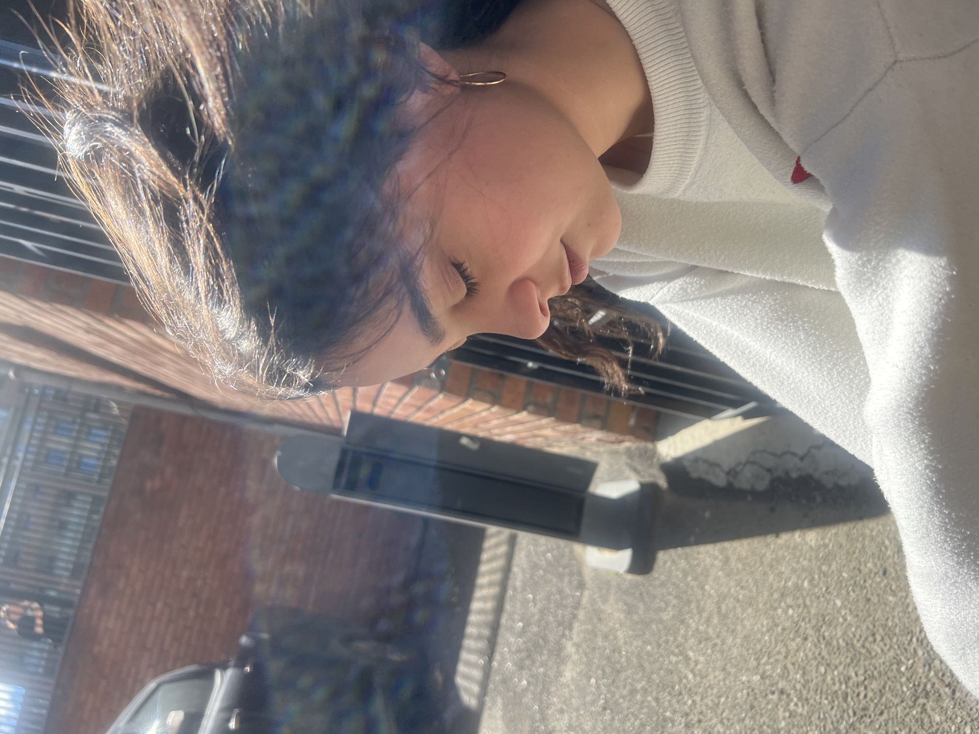 A young woman with dark hair and an earring stands outside a building in Tbilisi, Georgia. She is wearing a white sweater. A brick building is visible in the background along with a black car and gray pavement. There is a black metal grate near her. The image is rotated counterclockwise.