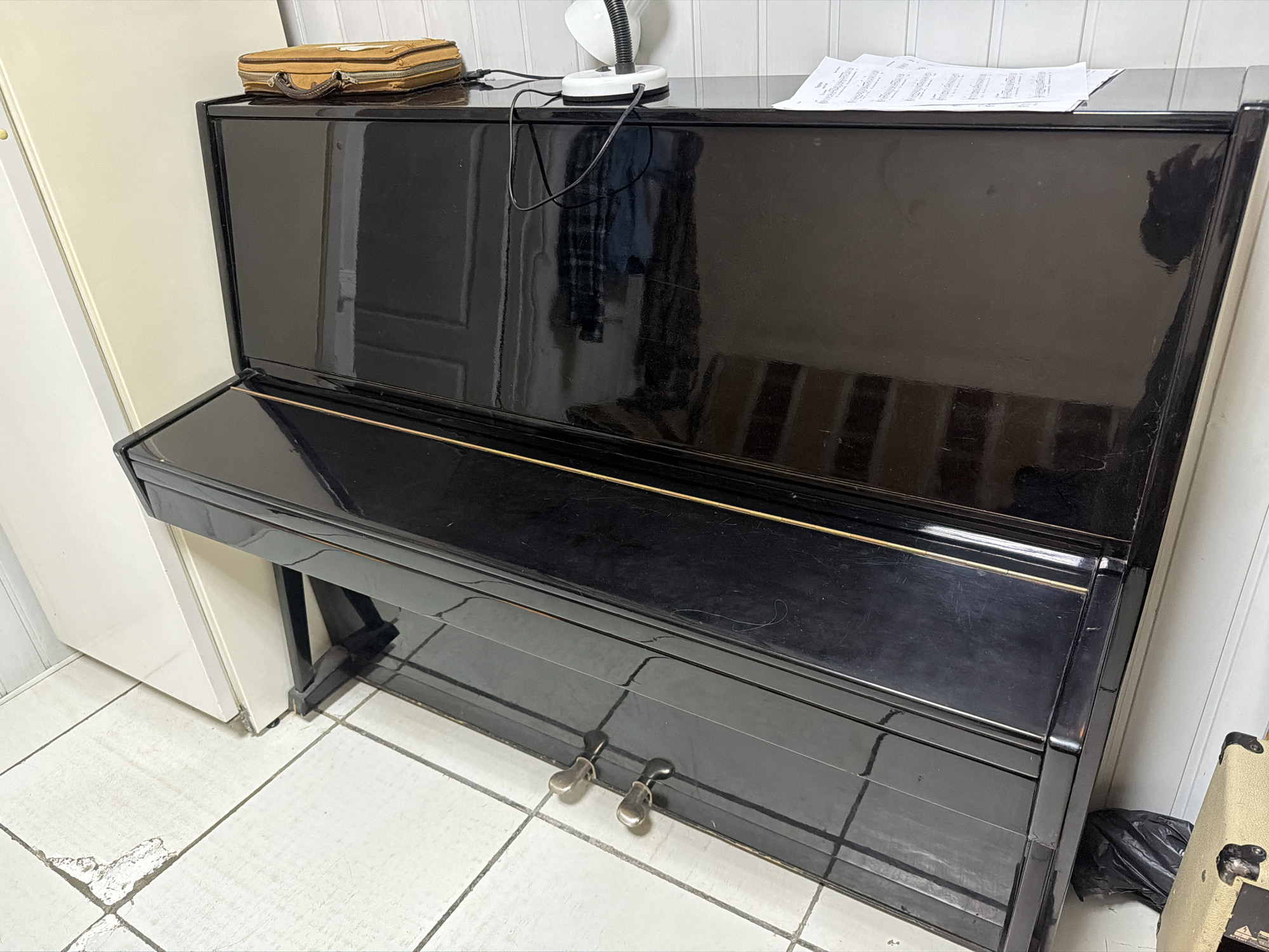 An eye-level shot captures a glossy black upright piano situated indoors. The piano is the central focus, showcasing its dark, reflective surface that mirrors aspects of the surrounding room, including what appears to be a door or window on its main panel.
On the top surface of the piano, several items are placed: to the left, a small, worn beige or light brown zippered case or pouch rests. Towards the center, a white desk lamp with a flexible black neck stands, connected by an electrical cord that trails across the piano's top. To the right, several sheets of music or paper are laid out, partially overlapping. The music sheets display musical notation and some faint, illegible text.
At the base of the piano, two silver-colored pedals are visible, indicating it is an acoustic piano. The floor beneath the piano consists of light-colored square tiles with dark grout lines. Some tiles on the left side show wear or damage, with portions chipped away.
To the immediate left of the piano, a section of a light-colored cabinet or door is visible. On the right side, a cream-colored amplifier or speaker with visible grilles and controls is partially seen, and a crumpled black plastic bag rests on the floor next to the piano. The background features light-colored, possibly paneled, walls. No human figures are present, and no specific activity is taking place. The lighting suggests an indoor setting, but the time of day is not discernible.