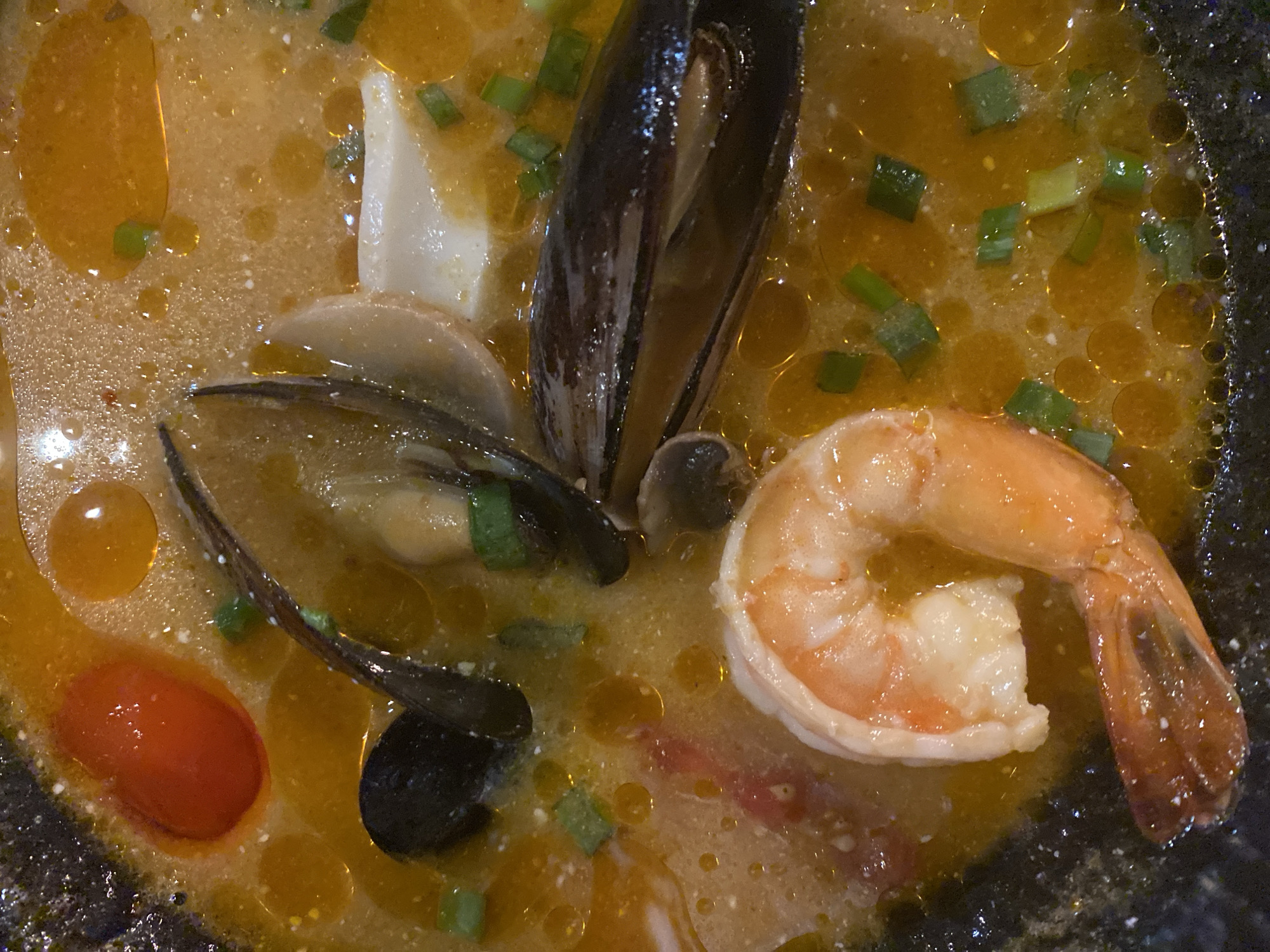 A close-up shot depicts a bowl of seafood soup. The soup contains mussels, shrimp, squid, and a tomato. Green onions are sprinkled on top. The soup is in a dark bowl, and there are oil droplets on the surface. The photo does not allow for a determination of Yekaterinburg, Russia.