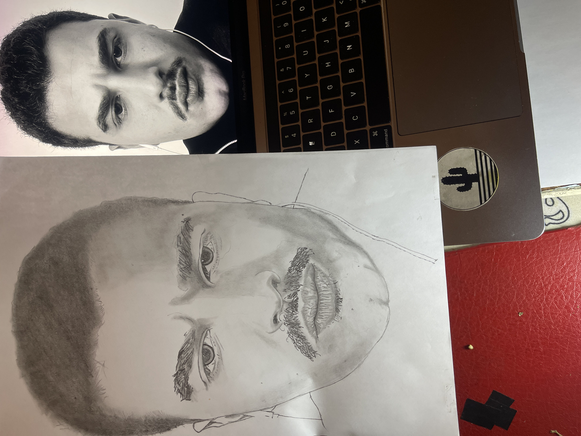 The image displays an indoor tabletop scene featuring a pencil drawing and a digital reference. On the left, a detailed graphite pencil drawing of a male face is rendered on white paper. The drawing depicts a man with short, dark hair, thick eyebrows, dark eyes, a handlebar mustache, and a slightly furrowed brow. To the right, an open dark-colored laptop, likely a MacBook Pro, displays a black and white photograph of the same male subject on its screen. In the photograph, the man wears white wired in-ear headphones and a dark zippered top, mirroring the expression of the drawing. A portion of the laptop's black QWERTY keyboard and trackpad is visible, along with a circular sticker on the palm rest featuring a black cactus silhouette. The surface below these items includes a textured red area on the left, containing two small black rectangular objects and two light-colored specks. A light-colored surface is visible beneath the laptop on the right, with a small section of what appears to be patterned currency at the bottom edge. The image provides no discernible visual information to identify its specific location within the city of Jos, Nigeria.
