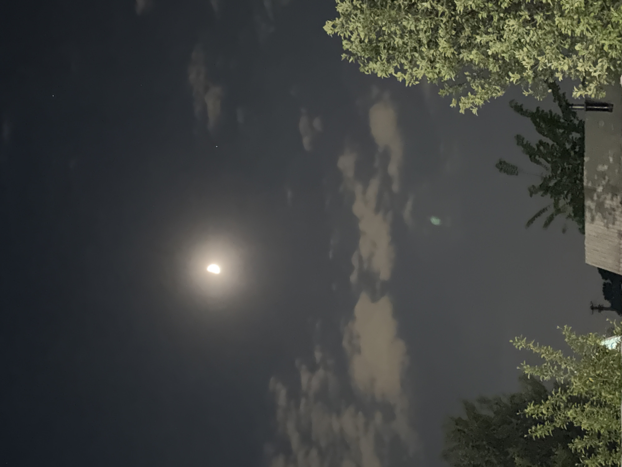 Here is a description of the image:
The image appears to be a nighttime shot of the sky with the moon visible. The moon is not full, showing only a crescent shape with a bright glow around it. There are light clouds partially obscuring the night sky. The bottom and right corners of the image show trees, and some architecture, including what appears to be a building with a pole. The image is a perspective of someone looking up from ground level.