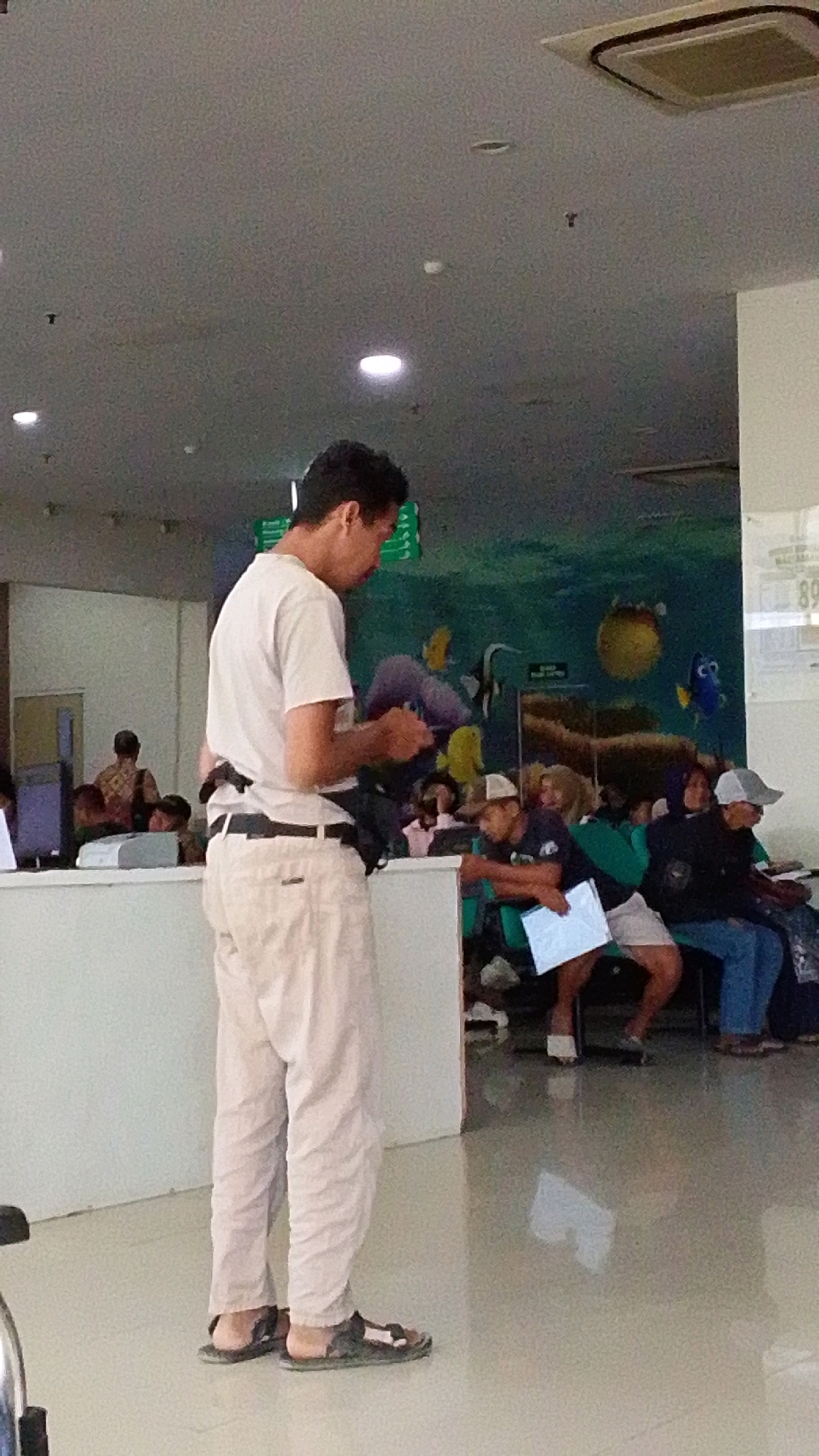 In a brightly lit indoor waiting area, possibly at a clinic or hospital in Pabelan, Indonesia, a man in light-colored pants and a white t-shirt stands facing away from the camera. He is holding a dark object in his hands and has a black bag slung over his shoulder. He is wearing sandals. Behind him, several other people are seated on green chairs, some holding papers. The wall behind them features a mural of underwater scenes with fish. The floor is tiled and reflective.