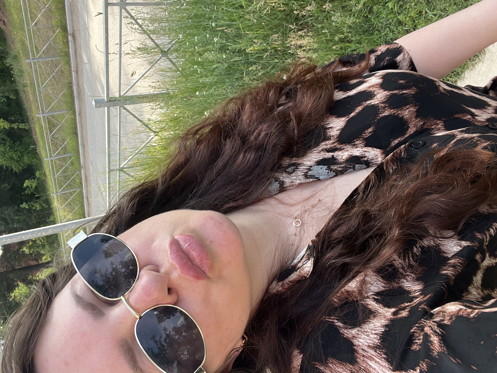A woman with long, dark, wavy hair, wearing sunglasses and a leopard-print dress, pouts her lips for a selfie. She is outdoors, with a metal fence and grassy area in the background. The scene is set in the city of Noviy, Russia, possibly near a road or park. The lighting suggests it is daytime, with sunlight casting shadows.