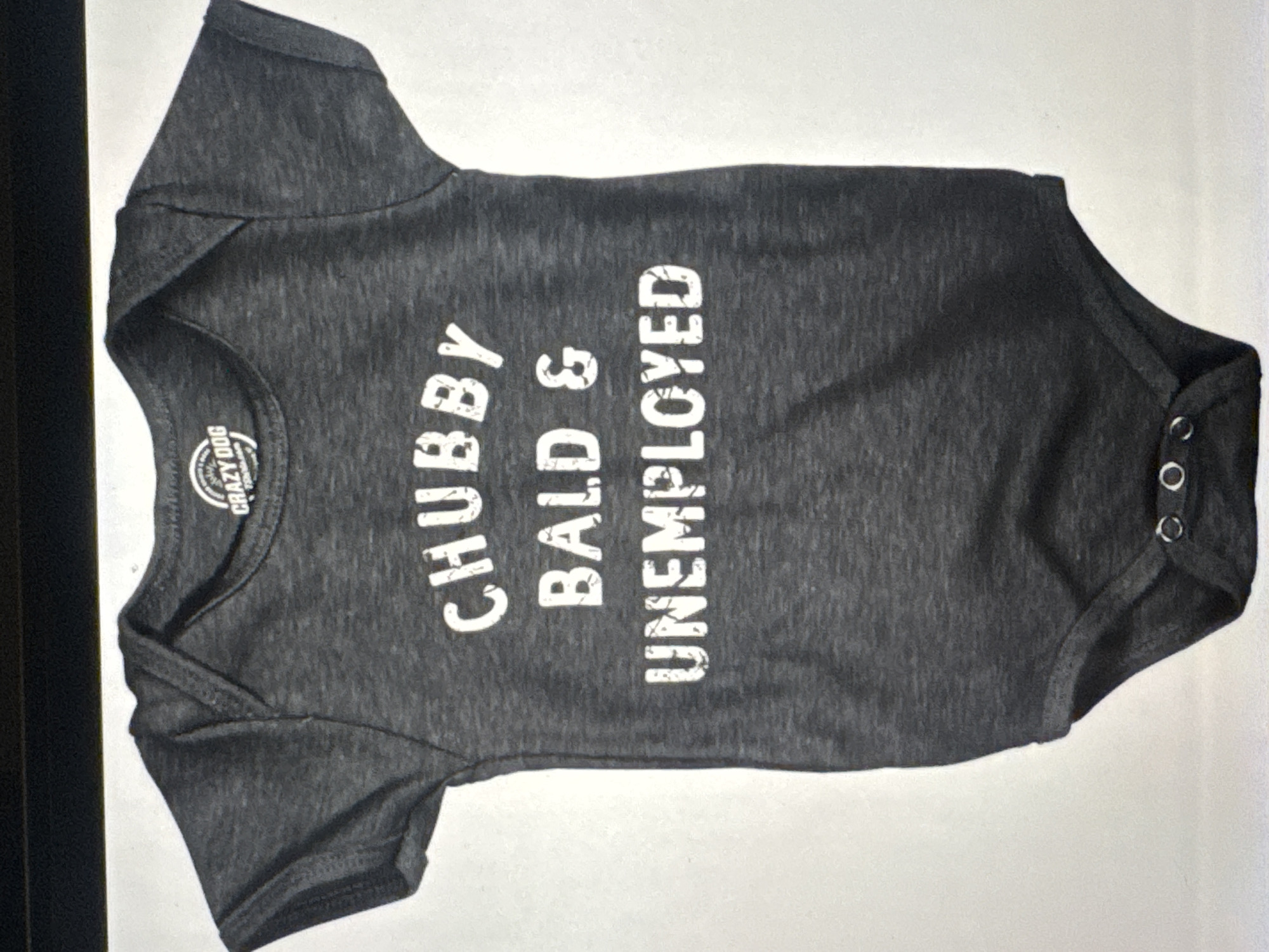 The image shows a close-up, high-angle shot of a dark gray baby onesie laid flat on a white surface. The onesie has white text printed on the chest in a distressed font. The text reads: "CHUBBY BALD & UNEMPLOYED". Above the text, on the collar, is a circular logo with text that is difficult to read clearly but appears to say "CRAZY DOG". The onesie has short sleeves and snaps at the bottom. The lighting is bright, casting minimal shadows. The background is plain white, with a dark horizontal line visible at the top of the frame, suggesting the edge of a table or shelf. There is no indication of the location in Kuta Selatan, Indonesia.