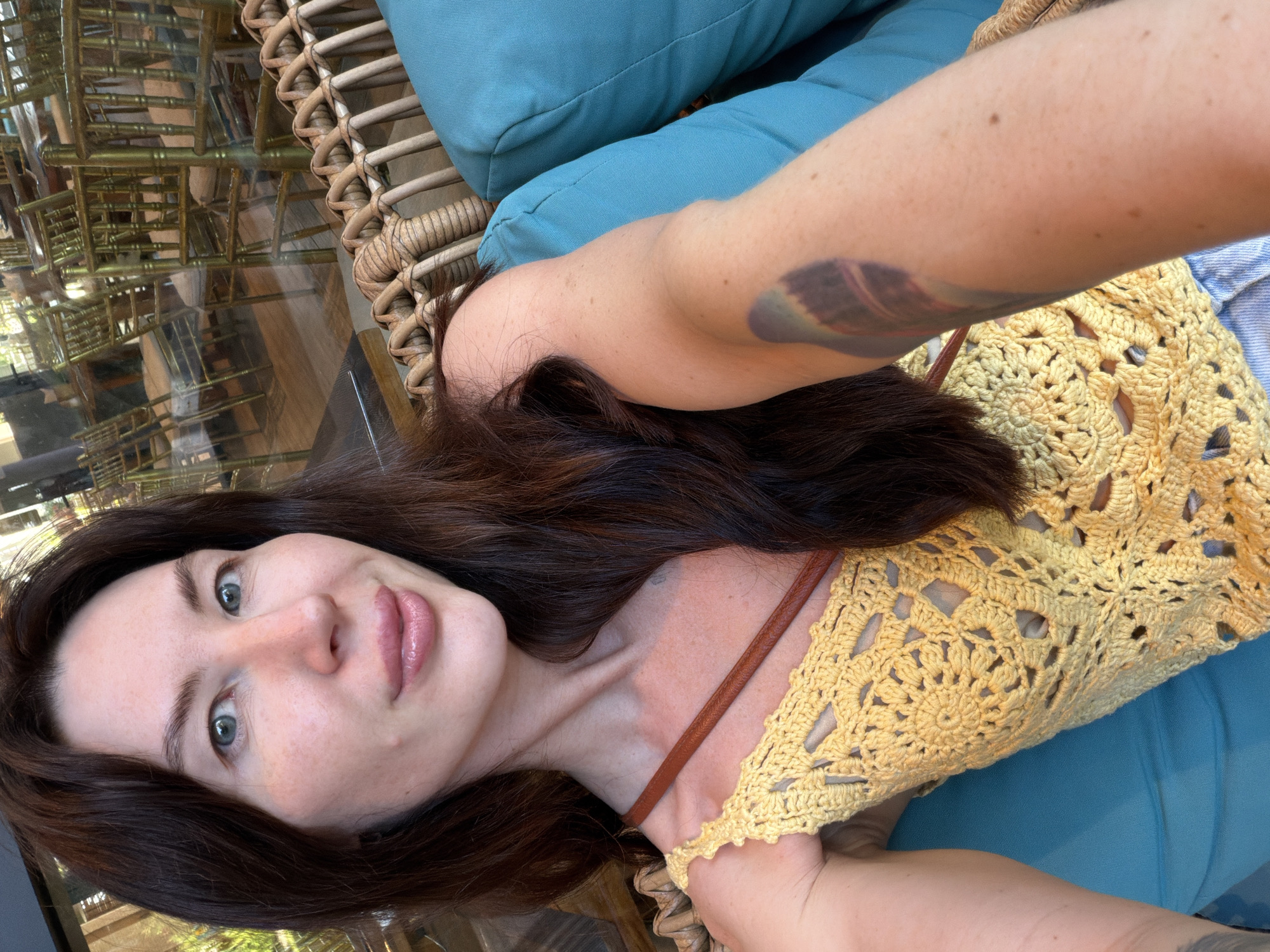 A woman with long, dark brown hair, fair skin, and light blue eyes is seated on a turquoise cushion, possibly in a restaurant or cafe. She is wearing a yellow crocheted top with a tan strap across her shoulder. Her left arm, visible in the foreground, has a tattoo. The background appears to be a blurred interior with golden chairs and tables, suggesting an indoor seating area. The scene is likely within a city, though its specific location in Dong Ninh Hoa Ward, Vietnam, is not explicitly depicted in the image itself, but rather a given context.