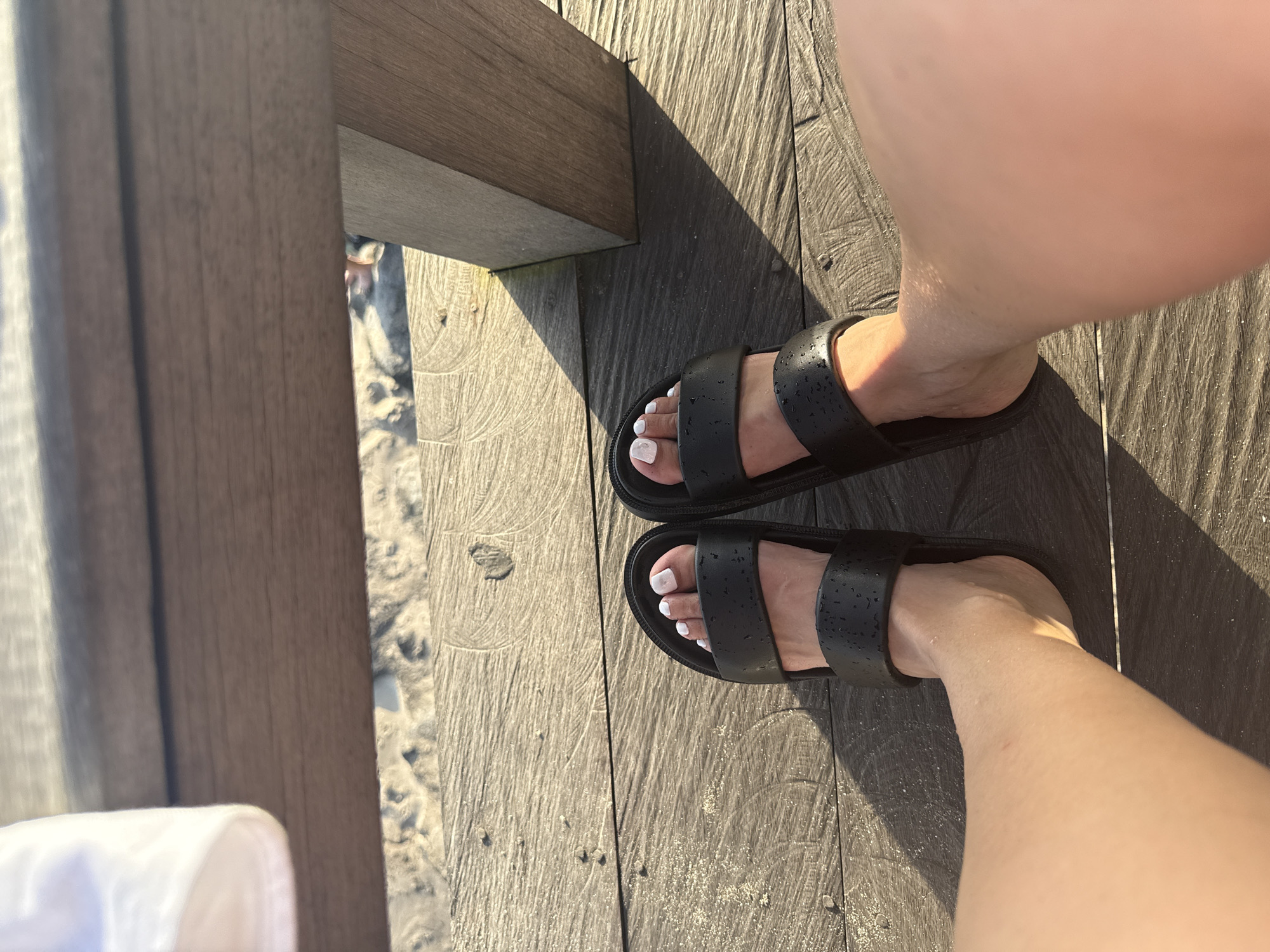 The image shows a person's feet in black sandals with white nail polish on a wooden deck. The deck appears to be adjacent to a beach, with sand visible between the wooden planks. The lighting suggests it is daytime, with shadows cast by the deck's support structure. Based on the style of the deck and beach setting, the location could be in North Kuta, Indonesia, known for its coastal resorts and beaches.