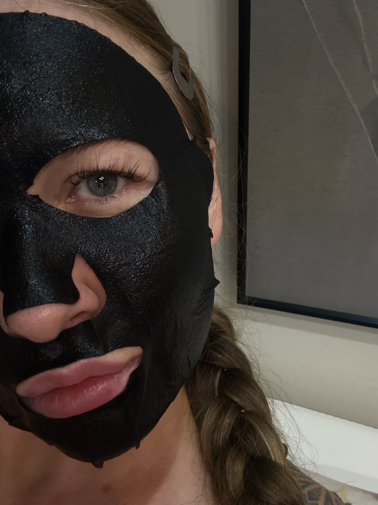 A close-up, head-and-shoulders shot shows the left side of a woman's face wearing a black charcoal sheet mask. The mask covers her entire face except for her left eye, nose, and mouth. Her eye is open and looking towards the camera, and her eyelashes are long and dark. Her lips are parted slightly, revealing a hint of teeth. Her hair is long and brown, braided into a single plait that falls over her right shoulder. A silver hair clip is visible near her temple. The background is out of focus, with a dark framed object on the right side of the image.