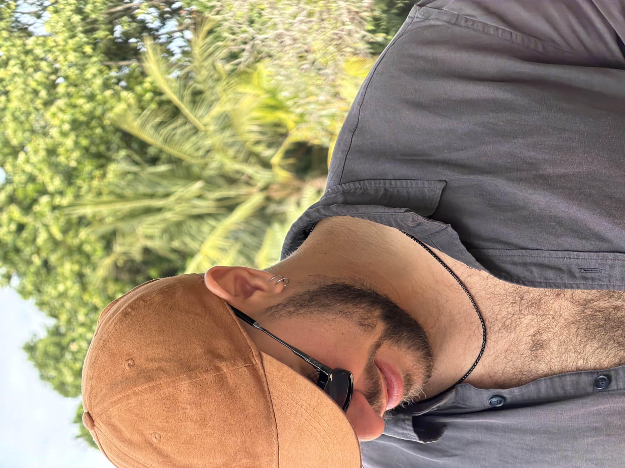 The image shows a close-up, outdoor shot of a man wearing a brown baseball cap, sunglasses, and a dark gray collared shirt. He has a beard and is wearing a silver chain necklace and hoop earrings. The background is blurred and consists of lush green foliage, likely trees and palm fronds, under a bright sky, suggesting a tropical or subtropical environment. The scene appears to be a casual, daytime setting, potentially in a garden or park area. While the specific location in Denpasar, Indonesia is not visually identifiable, the environment is consistent with the tropical climate found there.