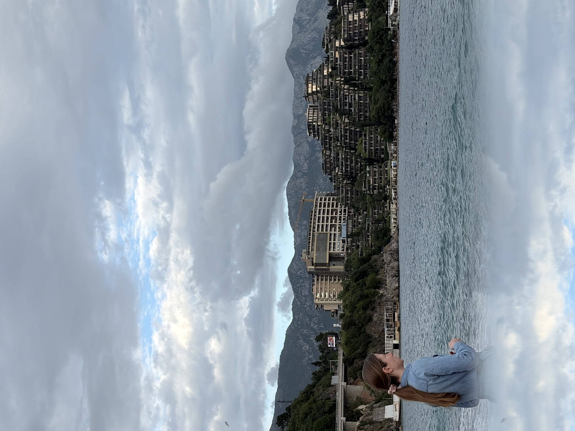 A horizontal, wide shot depicts a cloudy sky above water, with a tall landmass covered in buildings in the background and a woman looking toward the scene in the foreground. The woman is on the right, from the waist up, facing left. She has long brown hair in a ponytail and wears a denim jacket. She is on a rocky outcropping.
The water extends across the bottom half of the scene, with a shoreline of buildings and greenery beyond. The buildings are multi-level and appear to be apartments or hotels. Behind these buildings rises a steep, rocky mountain partially obscured by clouds.
The sky is overcast with varying shades of gray and white clouds. The image is taken during the day, and the light is diffused. The scene is Budva, Montenegro, known for its coastal resorts and architecture.