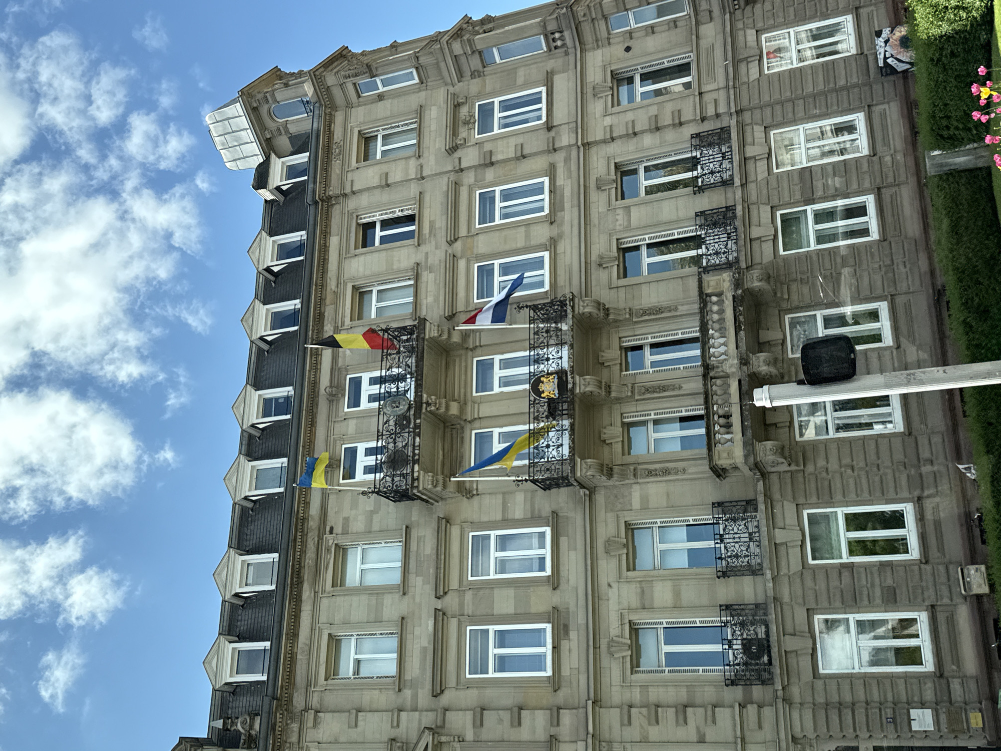 The image, rotated approximately 90 degrees counter-clockwise, presents an upward-looking view of the ornate stone facade of a multi-story building, set against a bright blue sky with scattered white clouds. The architecture, characteristic of classic European cities, features intricate decorative relief work, numerous rectangular windows with white frames (some with external blinds), and small wrought-iron balconies.
Multiple national flags are displayed from the building: a Belgian flag (black, yellow, red vertical stripes) and a French flag (blue, white, red vertical stripes) fly from two balconies. A Ukrainian flag (blue over yellow horizontal stripes) is visible higher up, near the roofline, and another appears lower down. One central balcony features a dark crest or emblem, though its details are indistinguishable. The upper part of the building has a dark-tiled roof with several dormer windows.
To the right of the building, a meticulously trimmed green hedge with small pink flowers adds a touch of nature. A white pole or fixture extends from the side of the building, and a small black, speaker-like object is visible below it. The clear sky and bright illumination confirm it is daytime. No people or specific events are depicted, focusing instead on the architectural grandeur and the prominent display of international flags, suggestive of a diplomatic, international, or historically significant establishment in Strasbourg, France. A small sign is affixed to the lower right part of the facade, but its text is unreadable.