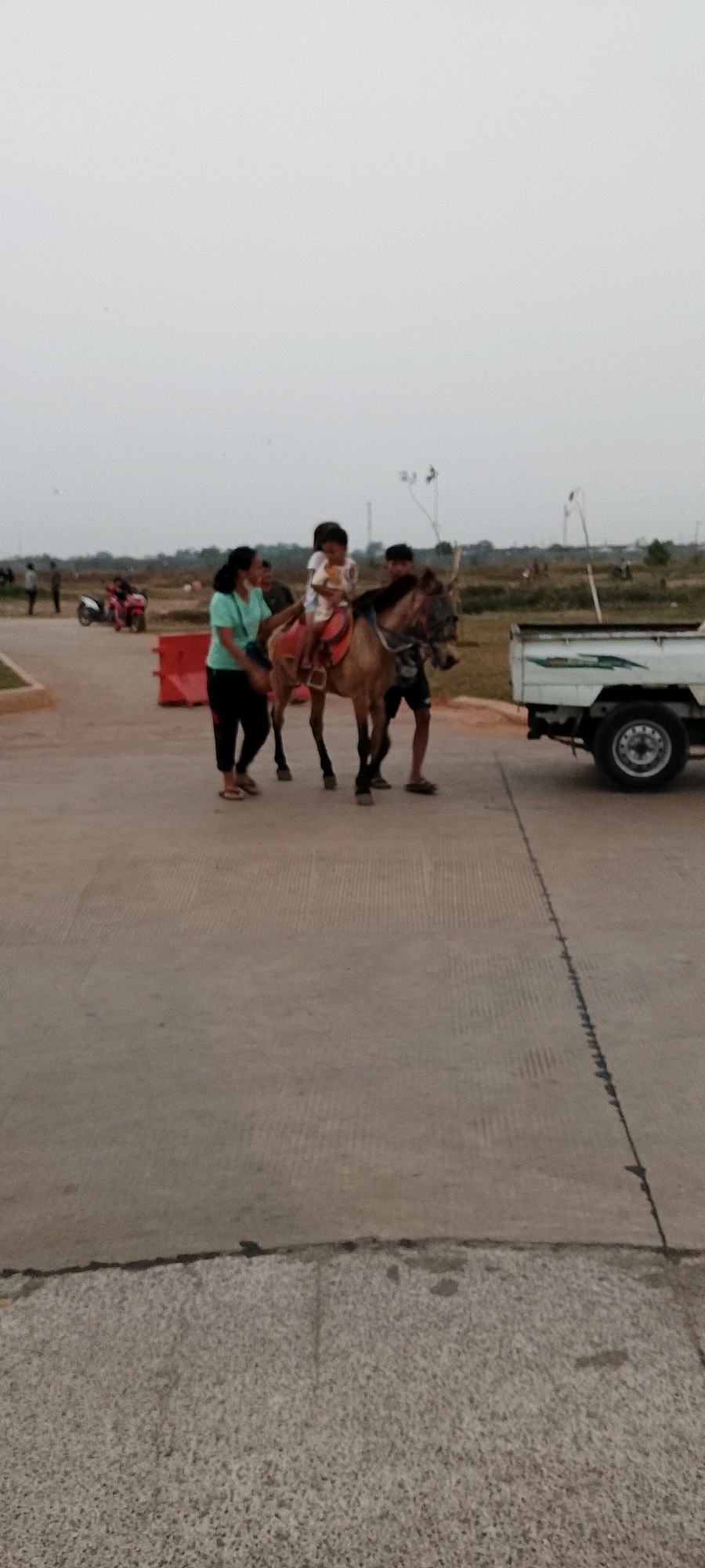 In the city of Satriamekar, Indonesia, a donkey is being led on a paved pathway. A young child is seated on the donkey's back, and an adult female in a teal shirt and black pants walks beside it, appearing to guide it. Another adult male, wearing dark shorts, walks behind the donkey. In the background, several motorcycles are parked, and a white pickup truck is visible on the right side of the frame. The environment suggests an outdoor, potentially recreational area, with flat, open terrain and a cloudy sky.