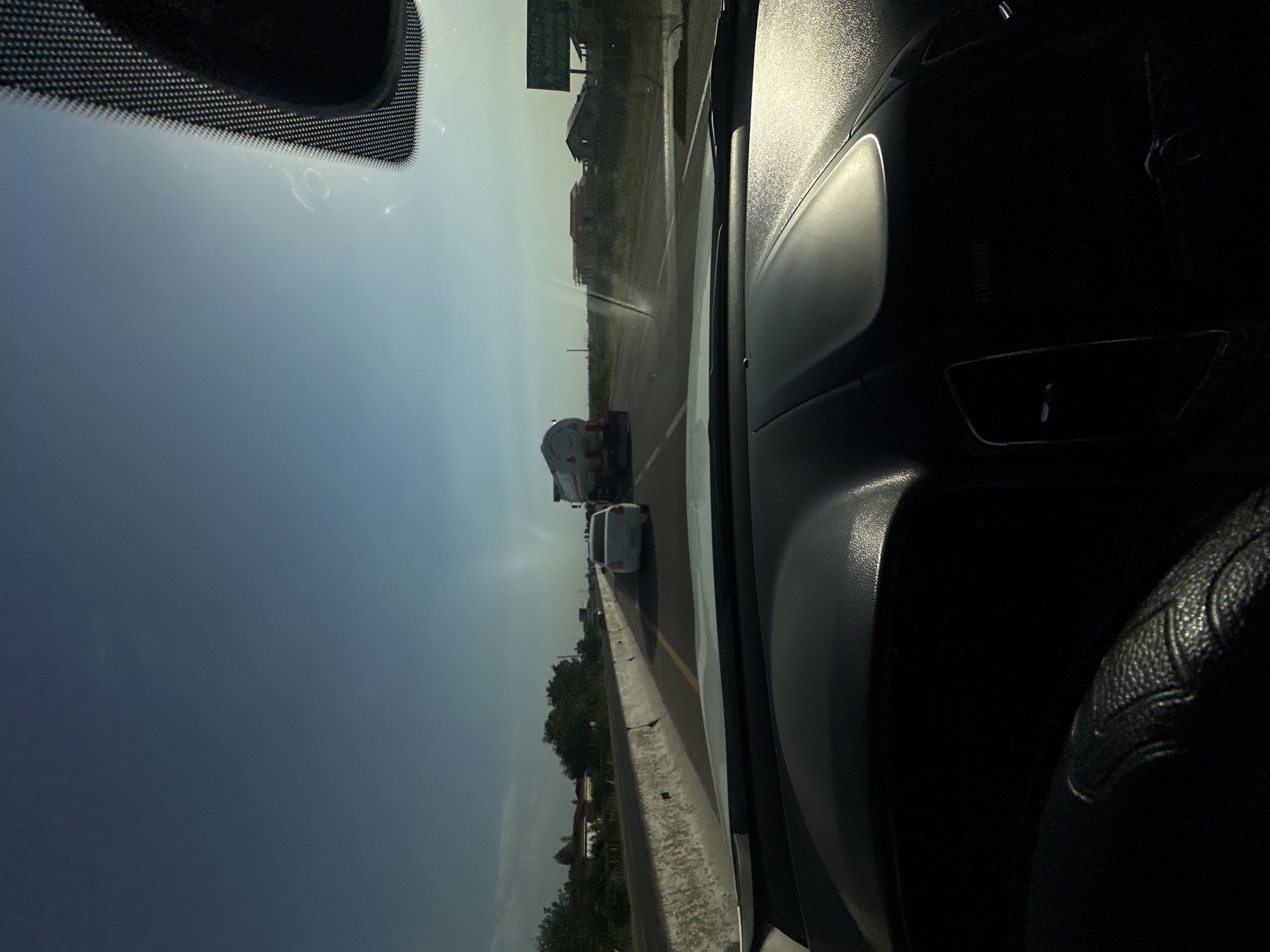 The image is taken from inside a vehicle, looking forward through the windshield. The sun is shining brightly, creating a glare on the windshield. The vehicle is on a highway, with other vehicles on the road. In the distance, there is a large tanker truck and a white car. The road is lined with a concrete barrier on the left and trees and buildings on the right. The sky is clear and blue. The interior of the vehicle, including the dashboard and steering wheel, is visible in the foreground. The image does not contain any information to suggest it is located in Tashtugizak, Uzbekistan.