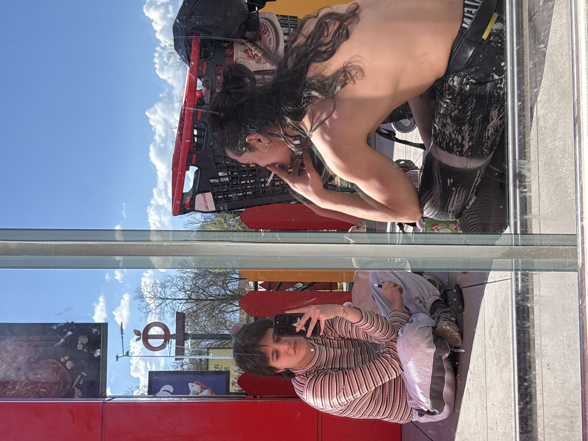 A reflection in a shop window shows two people. One individual, seen from the upper body up, is smoking a cigarette and is positioned near a red shopping cart. The other person is seated and taking a picture with a phone. The reflection includes a logo, and the environment suggests an outdoor urban setting, identified as the city of Sofia, Bulgaria, based on the logo visible in the reflection.