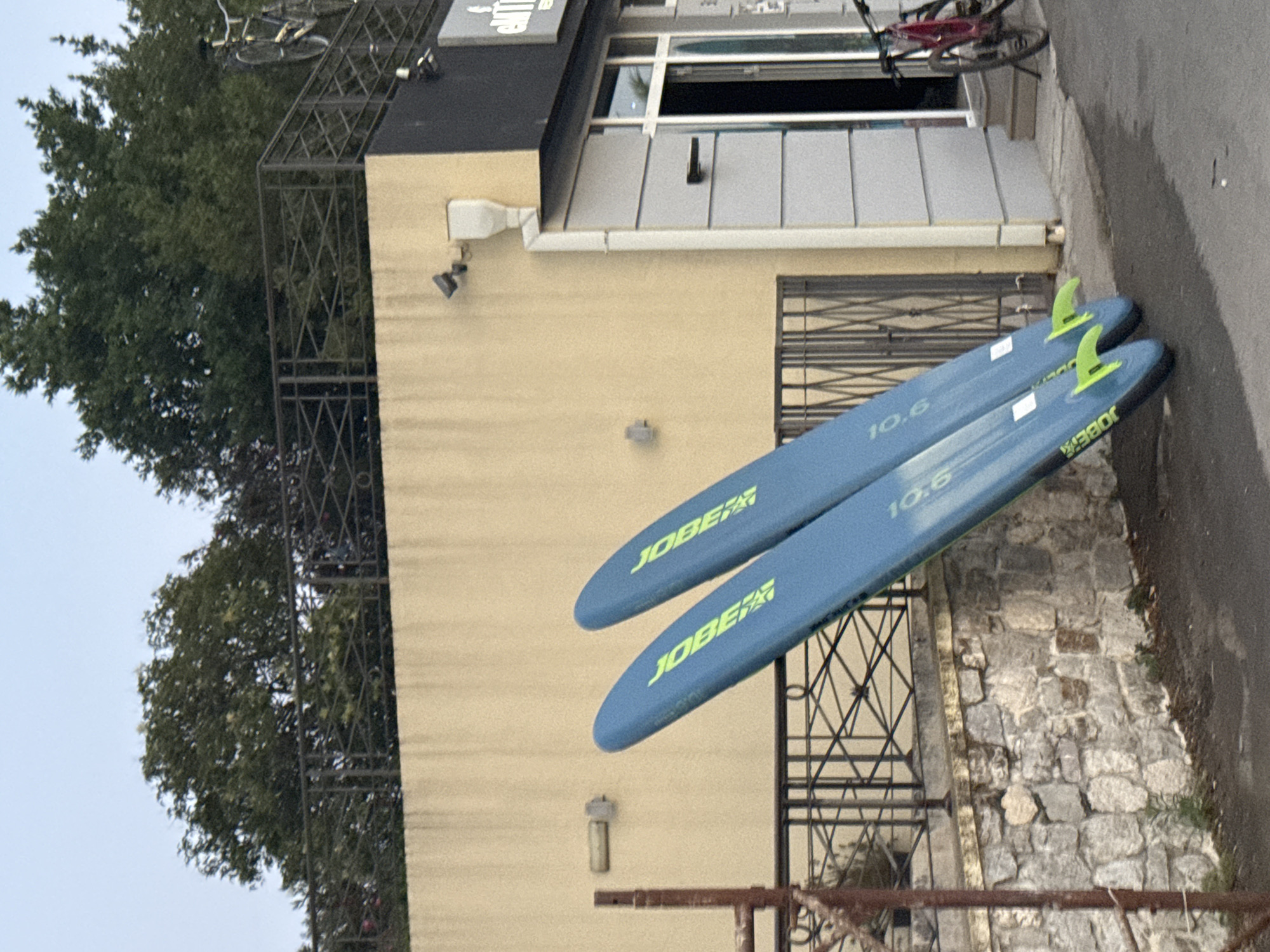 Two blue paddleboards with yellow fins and "JOBE" branding are leaning against a tan building. One paddleboard is positioned slightly in front of the other. The building has a dark awning and a silver-clad section with a doorway. To the right of the doorway, a red bicycle is parked. A stone wall and a metal railing are visible behind the paddleboards. Trees and a blue sky are in the background.