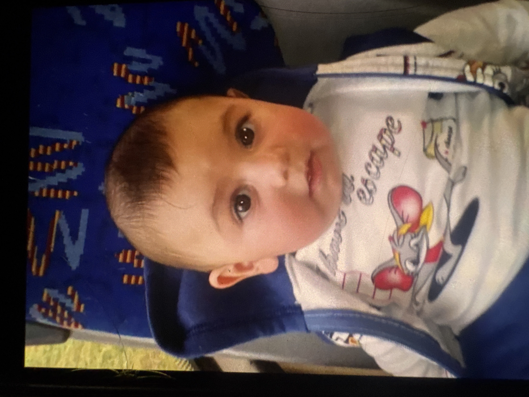 A close-up, slightly blurry, overhead shot shows a baby seated in a blue chair. The baby has brown hair and is wearing a white long-sleeved shirt with a picture of a cartoon mouse and the word "escape" on it. The background is a blue seat with a pattern of red and yellow shapes, possibly from a bus or train. The image is not taken in Rostov-on-Don, Russia, as no contextual elements suggest this location.
