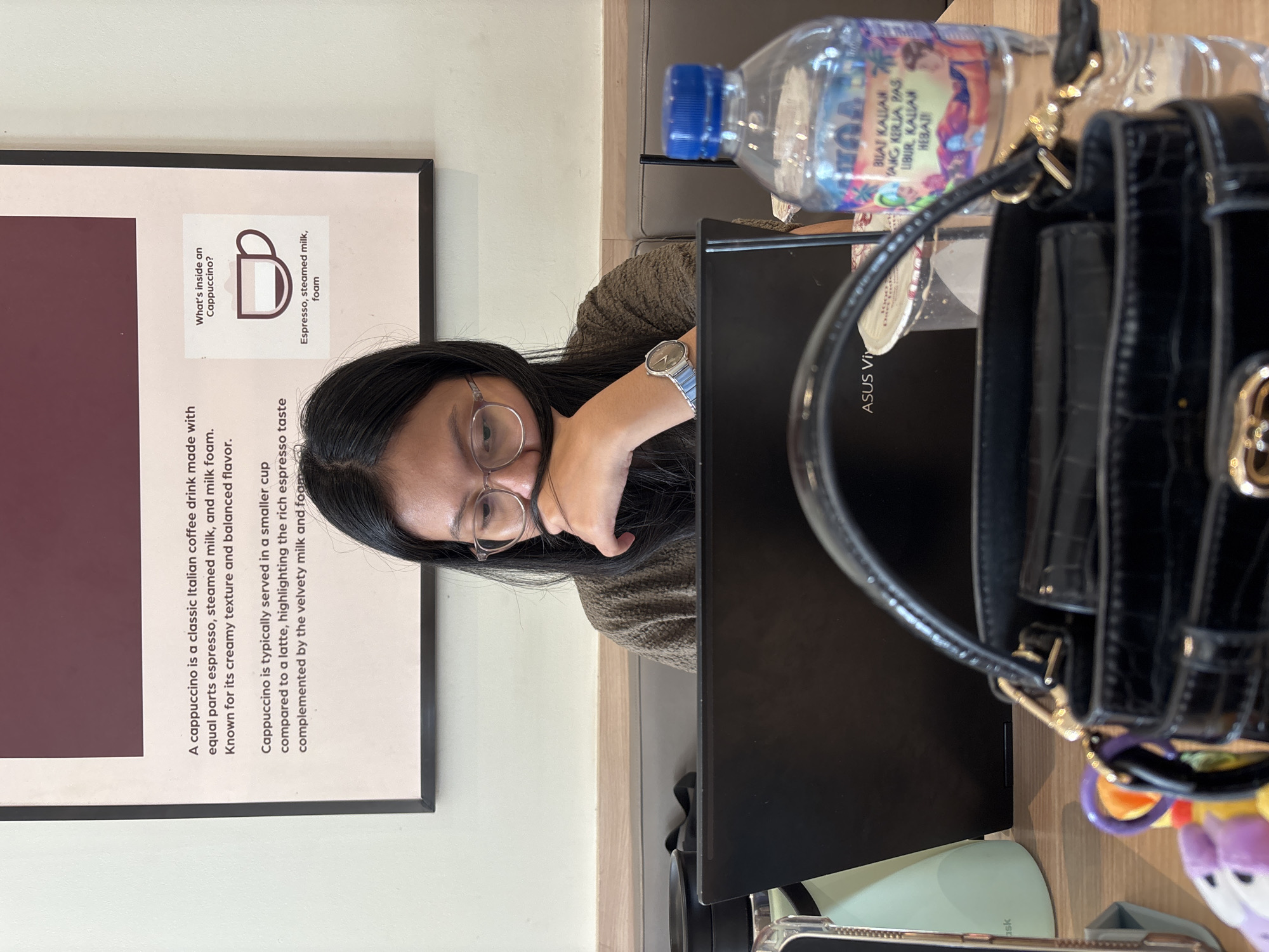 An Indonesian woman wearing glasses and a brown knitted sweater is sitting at a table, looking down at a laptop. Her hair is partially covering her face. In front of her is the laptop, a black handbag with a crocodile texture, and a partially visible plastic water bottle. Behind her, on the wall, is a framed poster with text describing a cappuccino. The environment appears to be a cafe or a similar establishment, possibly in Tangerang, Indonesia, as suggested by the presence of Indonesian text on the water bottle label.