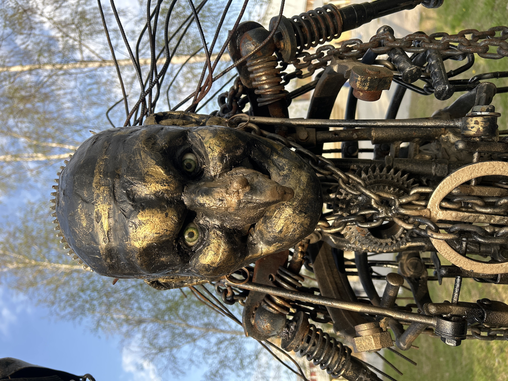The image depicts a metallic sculpture, likely a human head or face, with exaggerated features and a somber expression. The head is composed of dark, weathered metal with gold accents, suggesting rust or age. It's surrounded by a complex web of chains, gears, springs, bolts, and wires, all constructed from similar dark and weathered metal. These elements appear to be connected in a disorganized manner, giving the sculpture a mechanical or industrial aesthetic. The background shows sparse trees and a light blue sky, indicating an outdoor location. This artwork is located in the city of Uray, Russia.