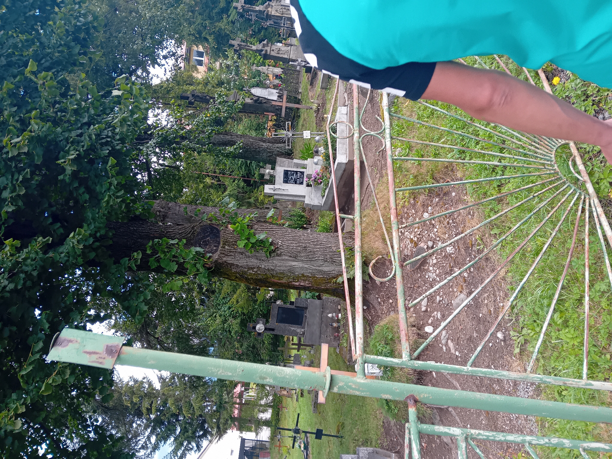 The image shows a cemetery in Zuberec, Slovakia. In the foreground, a person wearing a teal and black shirt is partially visible, facing away from the camera. A green, rusty metal gate with ornate detailing is in front of the person, obscuring the lower part of the cemetery. Beyond the gate, numerous gravestones and crosses of various sizes and materials are scattered across a grassy landscape. Tall trees with dense foliage provide shade over the burial ground. In the background, residential buildings and more trees are visible. The overall scene depicts a peaceful, albeit somber, burial site.