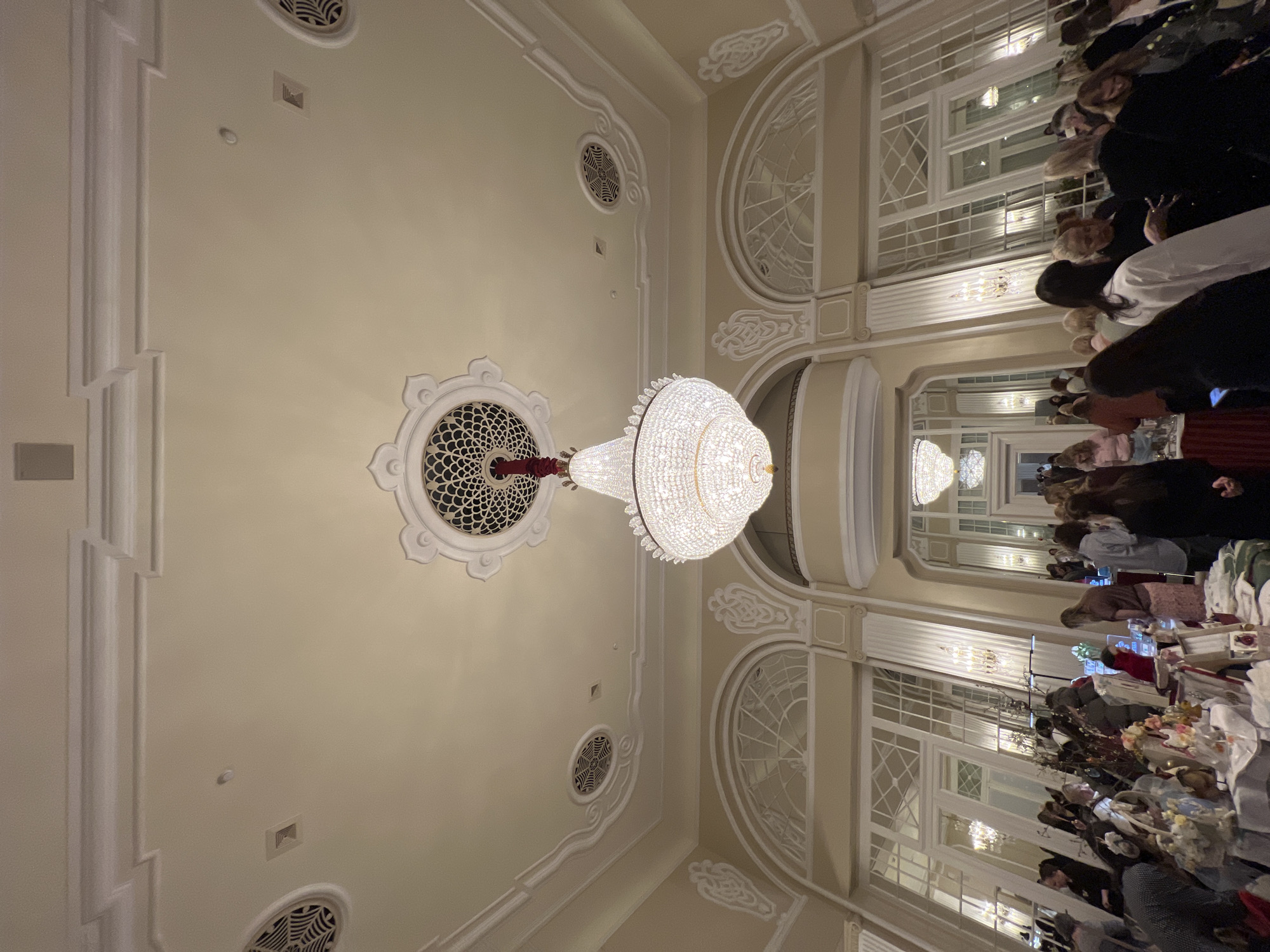 The image captures an ornate interior, likely a ballroom or grand hall, with a large, multi-tiered crystal chandelier as the central focal point. The ceiling is decorated with intricate plasterwork and circular vents. A series of arched windows line the right side of the frame, partially obscured by people and tables, suggesting an event or gathering in progress. The scene depicts a formal event, with numerous people in attendance, gathered around tables. It may be located in Saint Petersburg, Russia due to the architectural style.